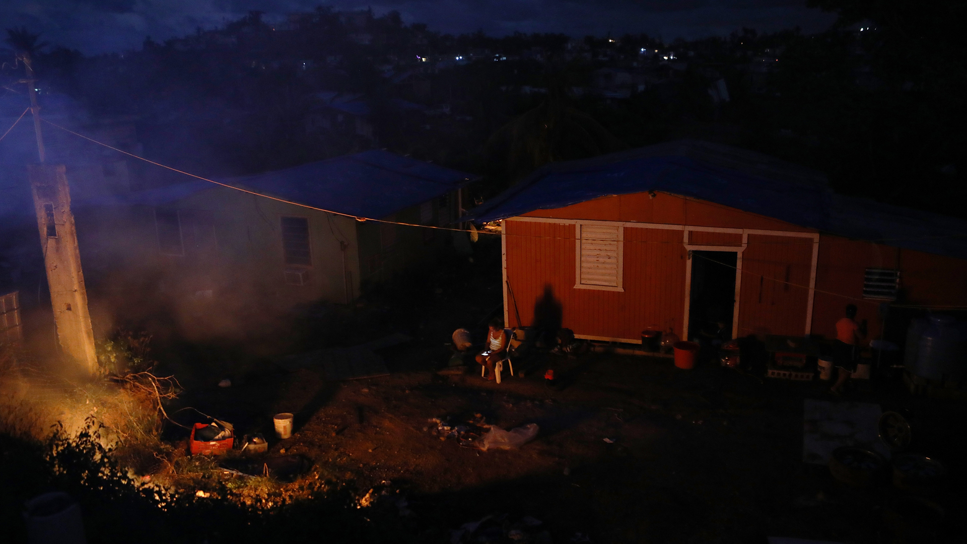 A resident sits outside her home, which lacks electricity, as a trash fire burns on December 21, 2017 in San Isidro, Puerto Rico. (Credit: Mario Tama/Getty Images)