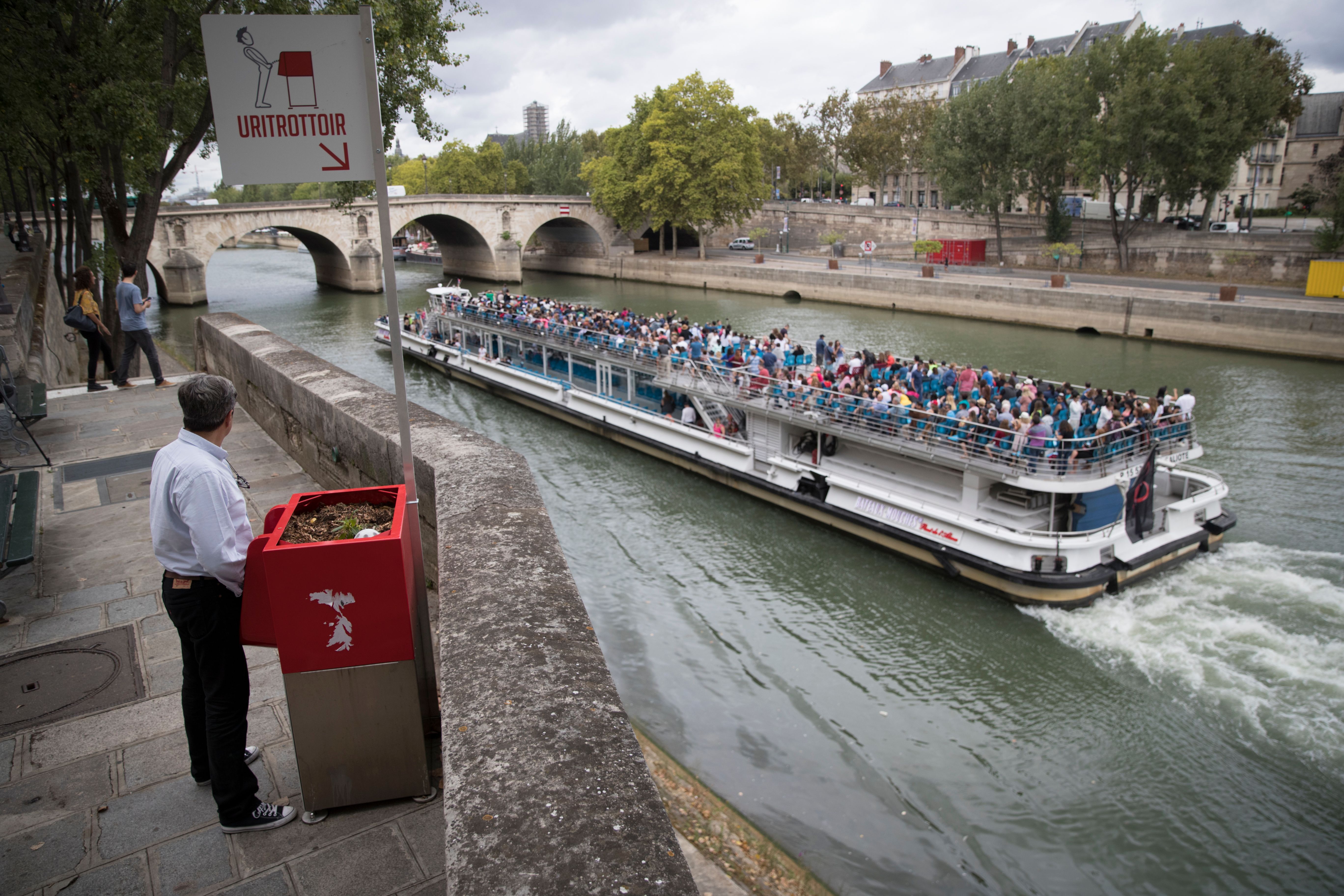 A man stands at a 'uritrottoir' public urinal on August 13, 2018, on the Saint-Louis island in Paris, as a 'bateau mouche' tourist barge cruises past. (Credit: THOMAS SAMSON/AFP/Getty Images)