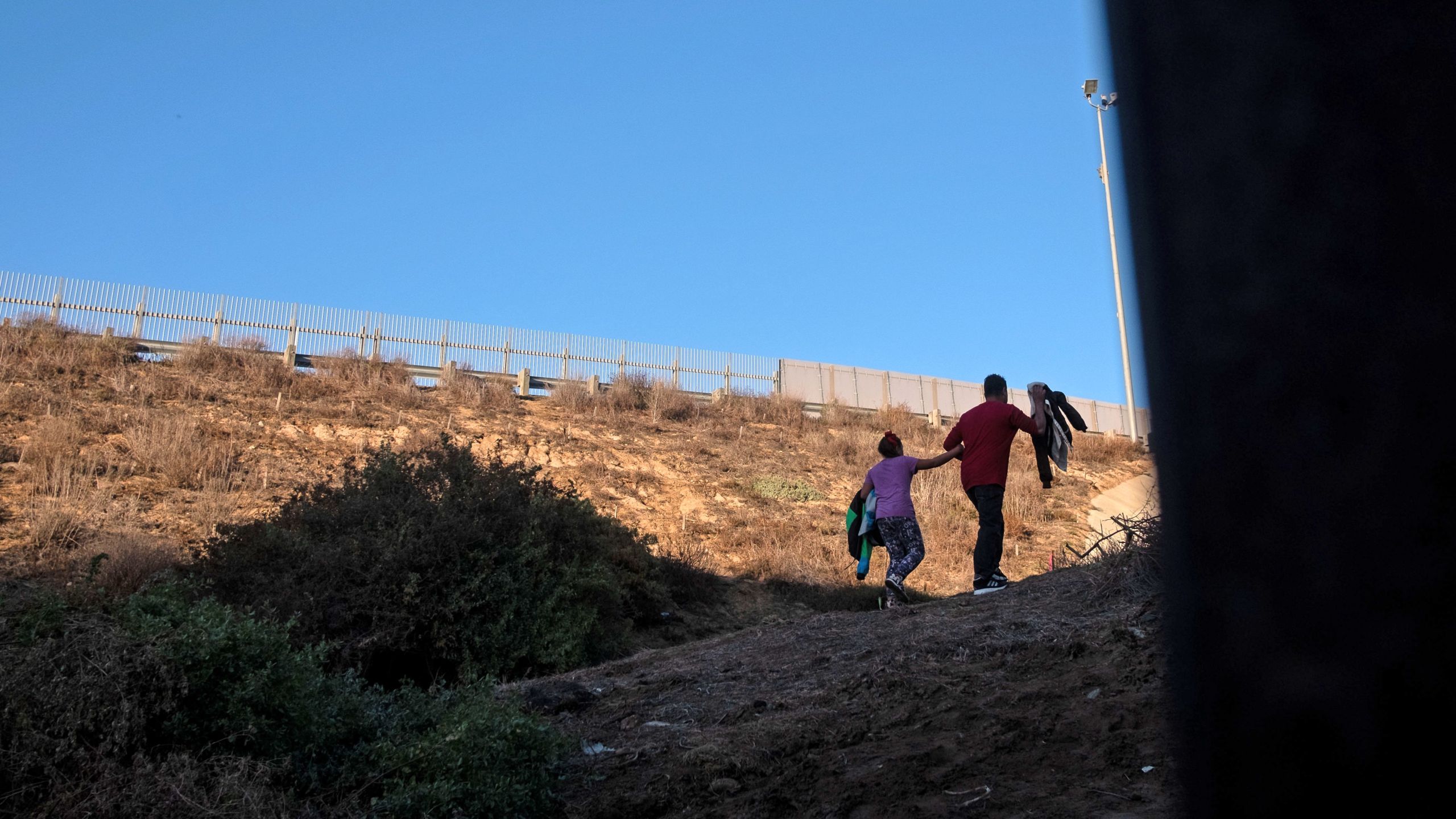 Honduran Andrea Nicolle, 10, and her father, Tony, traveling in a caravan of Central American migrants hoping to get to the United States, walk to surrender to the Border Patrol after crossing through a hole on the ground under the metal barrier separating Mexico and the U.S. on Dec. 4, 2018. (Credit: Guillermo Arias / AFP / Getty Images)