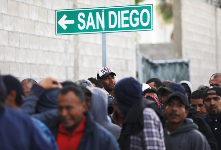 Migrants line up for free breakfast at the Desayunador Salesiano Padre Chava shelter and soup kitchen in front of sign for San Diego on March 9, 2018 in Tijuana, Mexico. (Credit: Mario Tama/Getty Images)