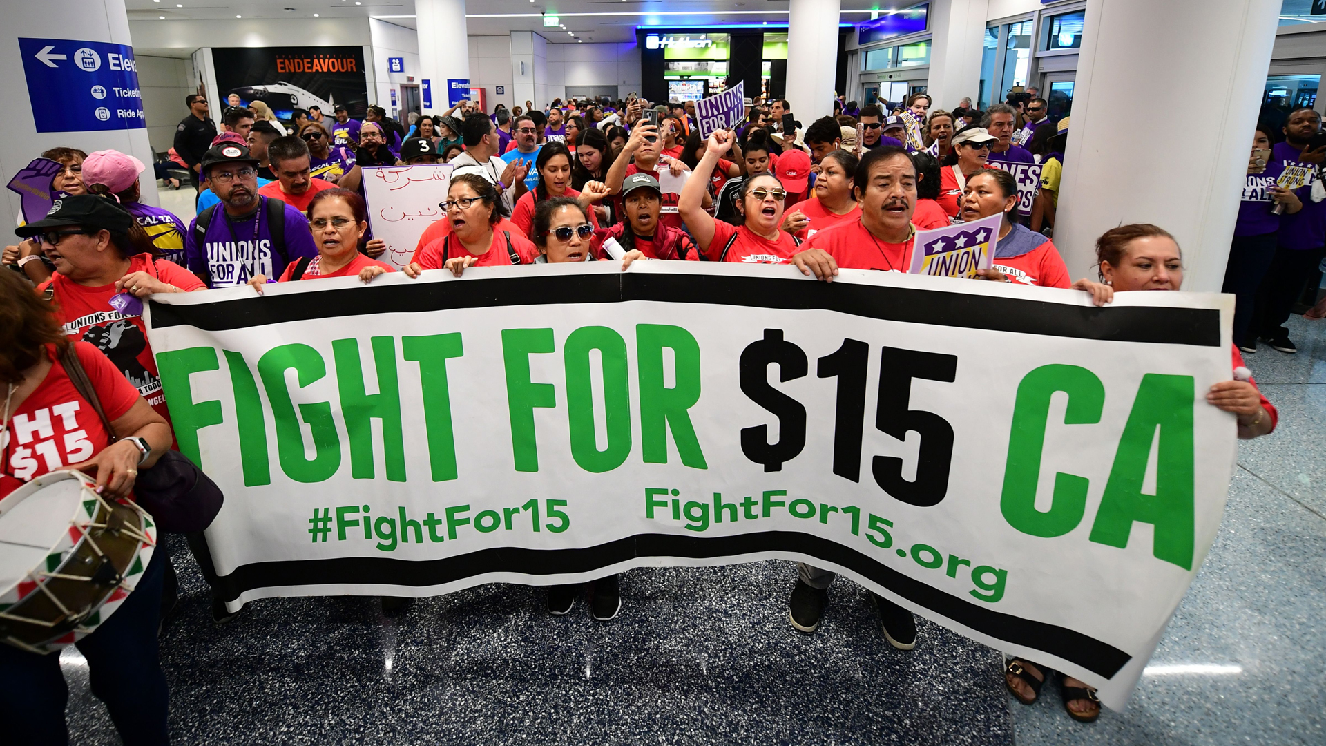 A banner for a $15 dollar per hour minimum wage is displayed as airport employees are joined by drivers from Uber and Lyft and other southern California workers for a protest at Los Angeles International Airport October 2, 2019. (Credit: FREDERIC J. BROWN/AFP via Getty Images)