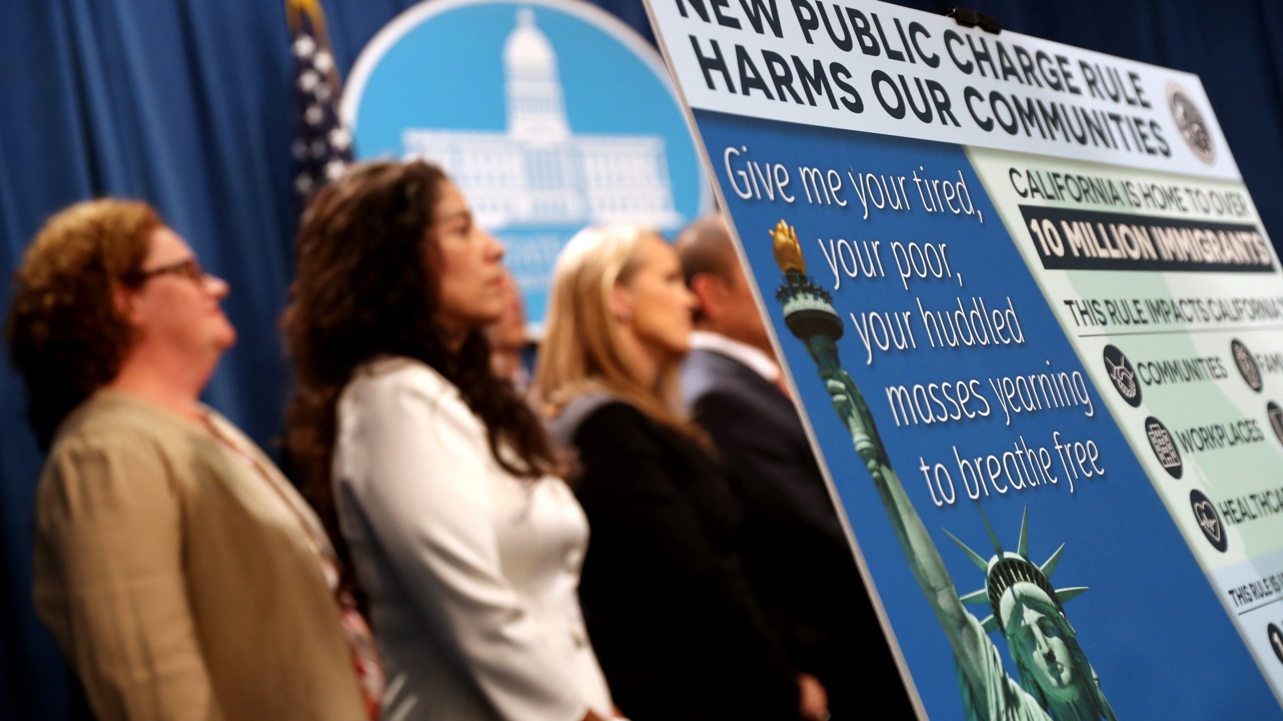 A sign is displayed during a news conference on Aug. 16, 2019 in Sacramento announcing a lawsuit against the Trump administration over the legality of a "public charge" rule that would make it difficult for immigrants on public assistance to obtain green cards. (Credit: Justin Sullivan/Getty Images)