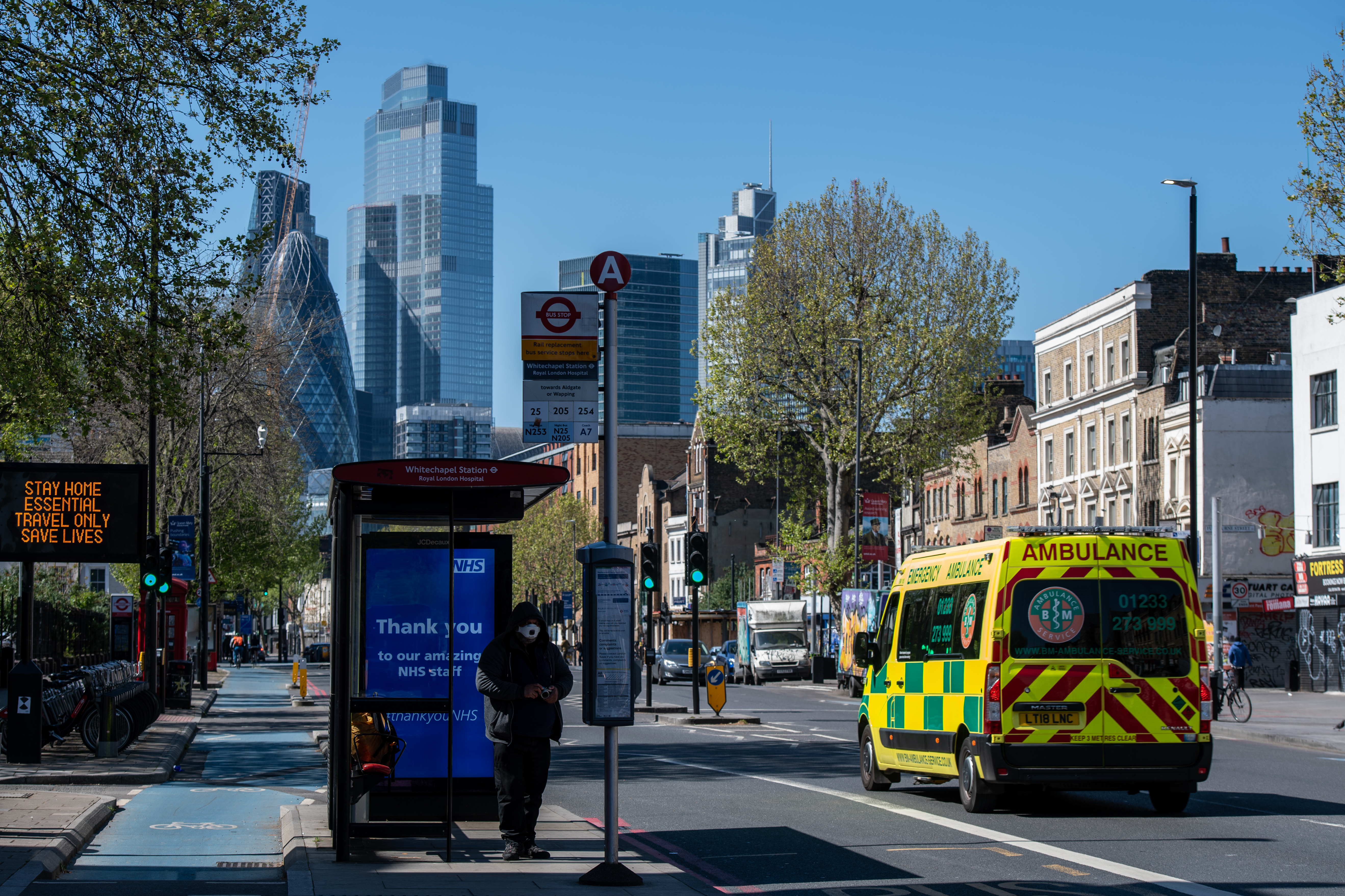 Skyscrapers are seen in the distance as a man wearing a face mask waits at a bus stop outside Royal London Hospital on April 19, 2020. (Chris J. Ratcliffe/Getty Images)