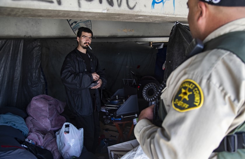 L.A. County sheriff’s Deputy Michael Tadrous of the Homeless Outreach Services Team talks with Shawn Troncozo, 24, about how to prevent the spread of the coronavirus near the San Gabriel River in this undated photo. (Credit: Gina Ferazzi / Los Angeles Times)