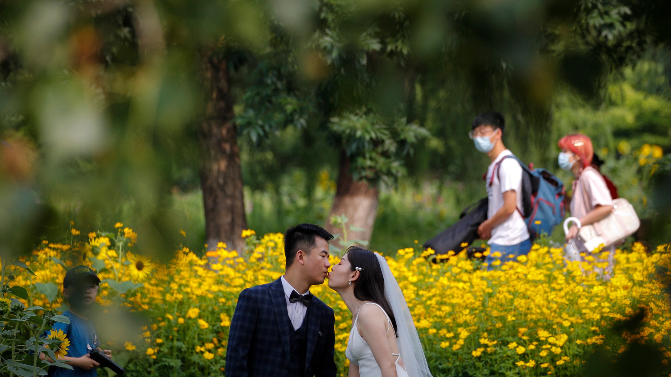 People wearing face masks pass by newlyweds kissing as they posing for wedding photos at the Olympic Forest Park in Beijing on July 2, 2020. (AP Photo/Andy Wong)