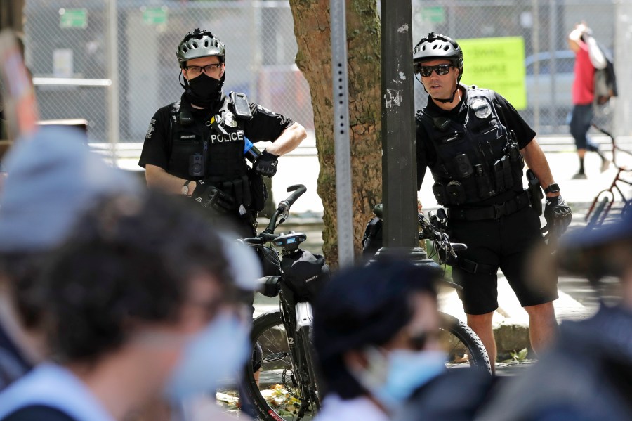 In this July 20, 2020, file photo, police officers look on at protesters in Seattle. (AP Photo/Elaine Thompson, File)