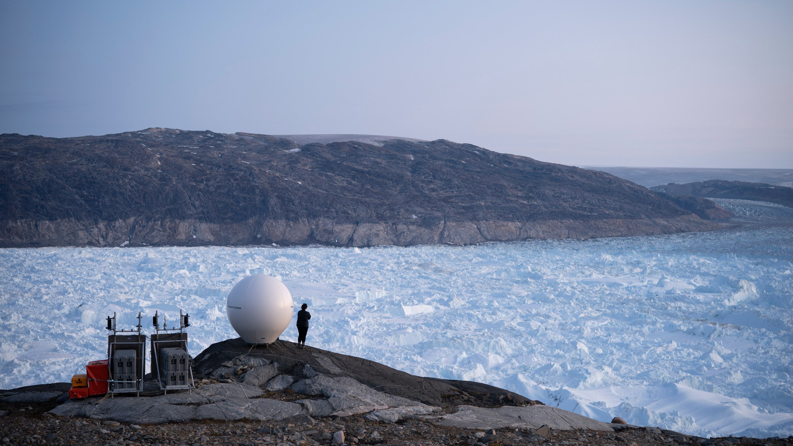 In this Aug. 16, 2019 file photo, a woman stands next to an antenna at an NYU base camp at the Helheim glacier in Greenland. According to a study released on Thursday, Aug. 20, 2020, Greenland lost a record amount of ice during an extra warm 2019, with the melt massive enough to cover California in more than four feet (1.25 meters) of water. (AP Photo/Felipe Dana)