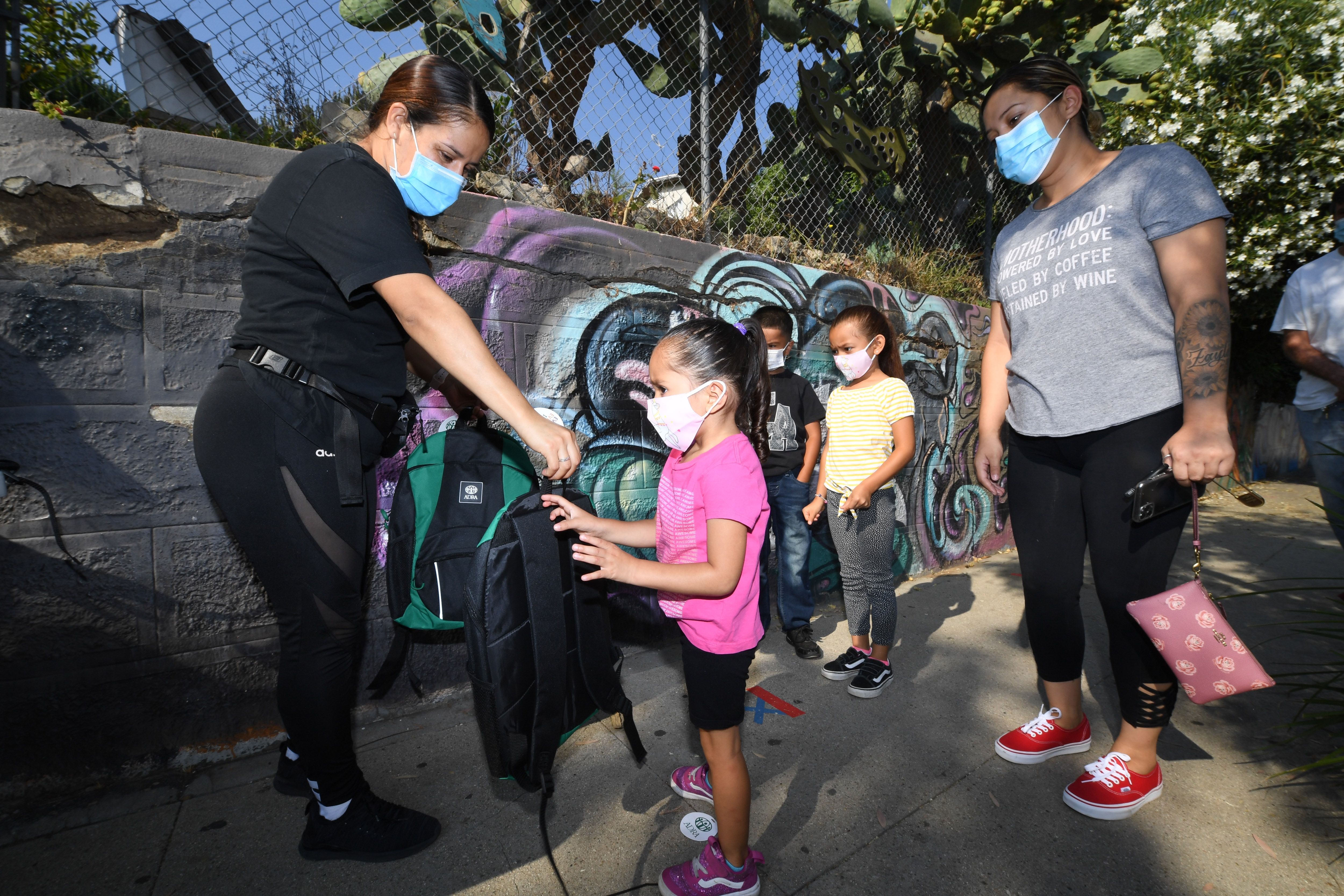 A volunteer hands 3-year-old Zayla a backpack filled with school supplies at a distribution to support neighborhood families as her mom Claudia Rodriguez, right, looks on, Aug. 14, 2020 in Los Angeles. (ROBYN BECK/AFP via Getty Images)