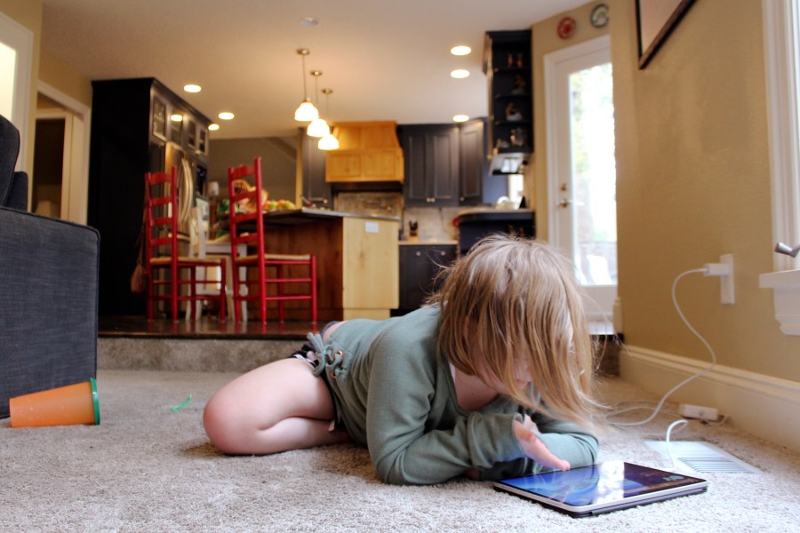 Lizzie Dale sprawls on the floor to play games on an iPad as her siblings work on school work in the kitchen behind her in their home in Lake Oswego, Ore., Oct. 30, 2020. (Sara Cline/AP Photo)