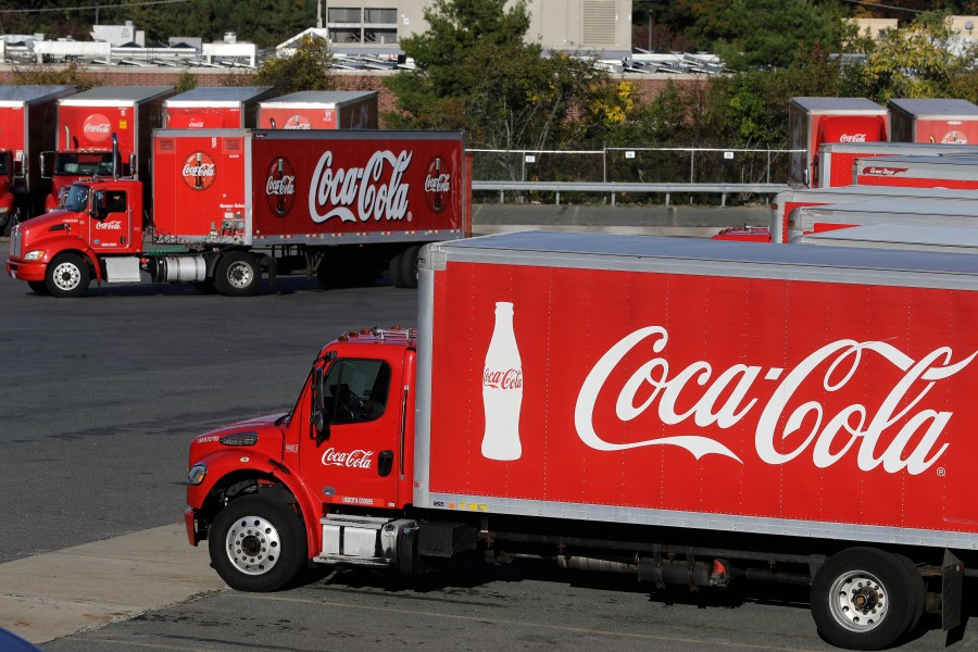 In this Oct. 14, 2019, photo a truck with the Coca-Cola logo, behind left, maneuvers in a parking lot at a bottling plant in Needham, Mass. (AP Photo/Steven Senne, File)