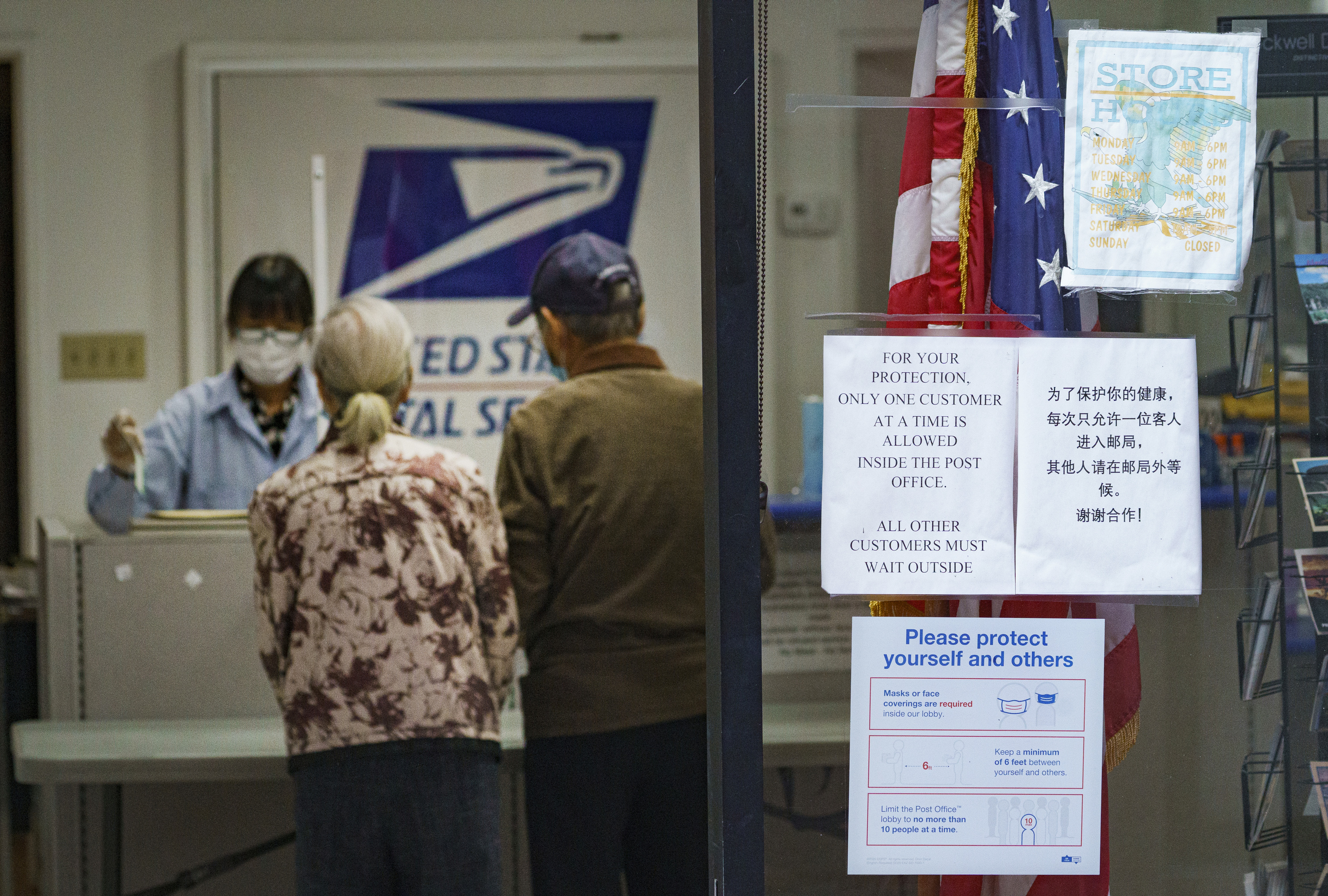 A sign posted at a U.S. post office advises that only one customer at a time is allowed inside, as a senior couple mails a parcel in the Chinatown district of Los Angeles Wednesday, Nov. 18, 2020, during the coronavirus pandemic. (AP Photo/Damian Dovarganes)