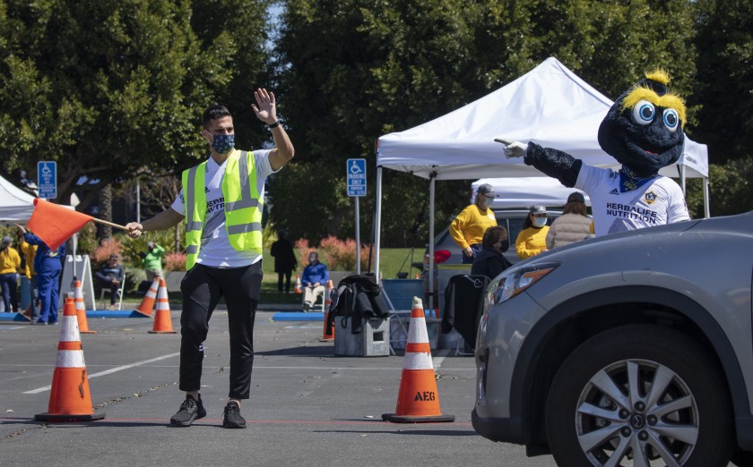 Galaxy midfielder Sebastian Lletget and team mascot Cozmo help direct traffic on March 16, 2021 ,at Dignity Health Sports Park in Carson, the home of Galaxy soccer, which was turned into a COVID-19 vaccination site. (Myung J. Chun / Los Angeles Times)