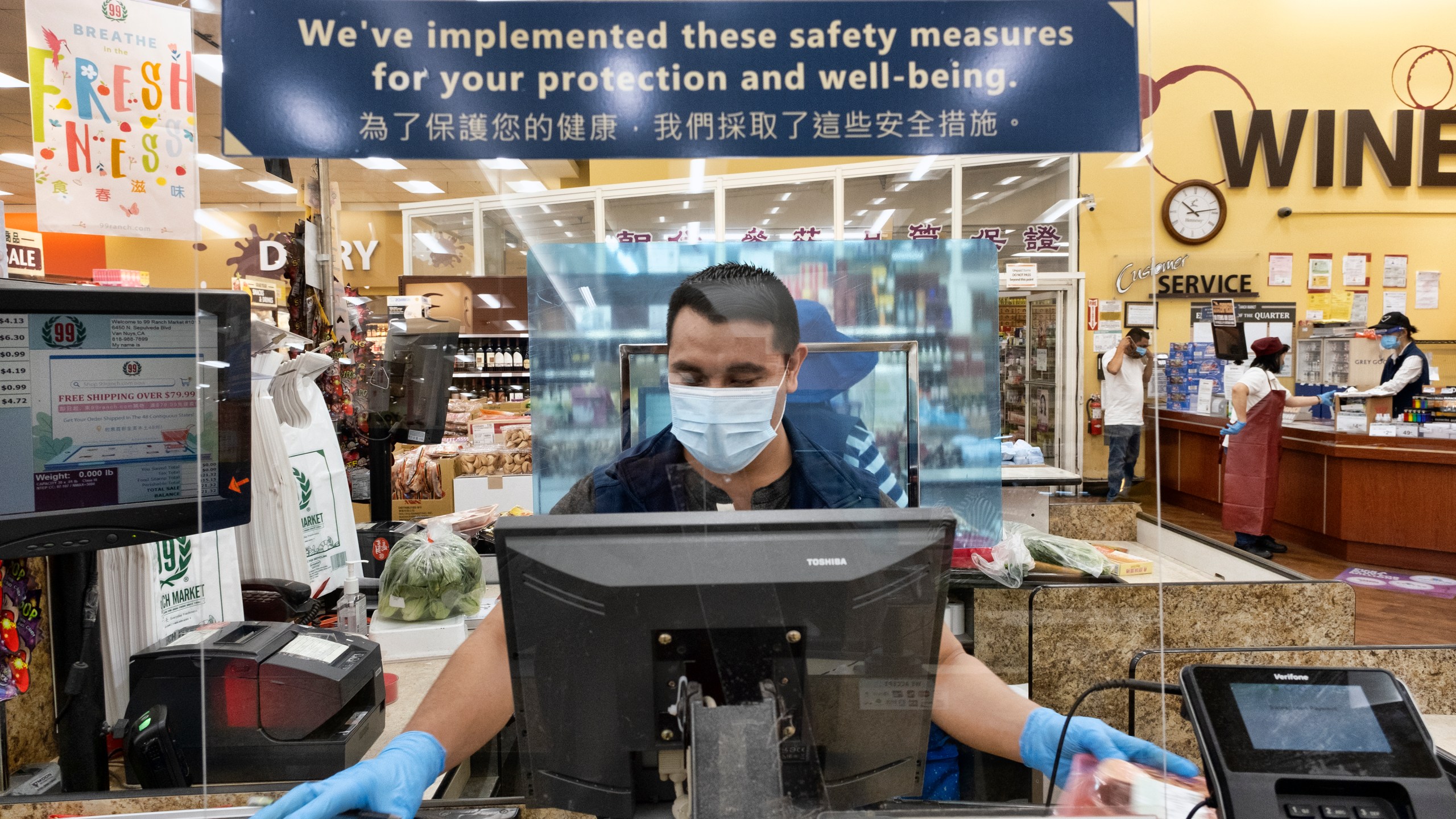 A grocery worker wears a protective mask and gloves as he helps to check out a customer from behind a plexiglass barrier at a market Van Nuys on May 5,2020. (Richard Vogel / Associated Press)