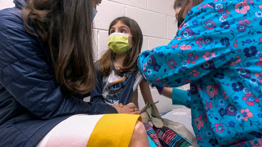 In this Wednesday, March 24, 2021 image from video provided by Duke Health, Alejandra Gerardo, 9, looks up to her mom, Dr. Susanna Naggie, as she gets the first of two Pfizer COVID-19 vaccinations during a clinical trial for children at Duke Health in Durham, N.C. In the U.S. and abroad, researchers are beginning to test younger and younger kids, to make sure the shots are safe and work for each age. (Shawn Rocco/Duke Health via AP)