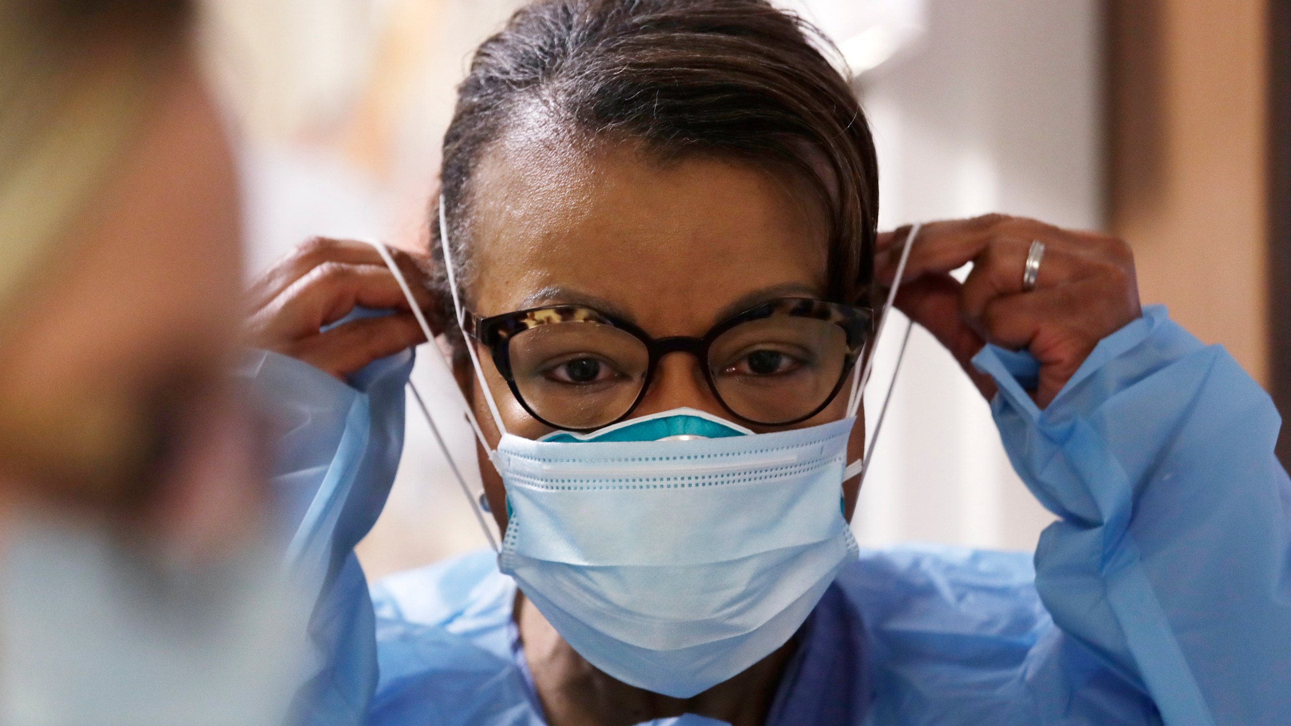 In this Friday, May 8, 2020 file photo, a respiratory therapist pulls on a second mask over her N95 mask before adding a face shield as she gets ready to go into a patient's room in the COVID-19 Intensive Care Unit at a hospital in Seattle. (AP Photo/Elaine Thompson)