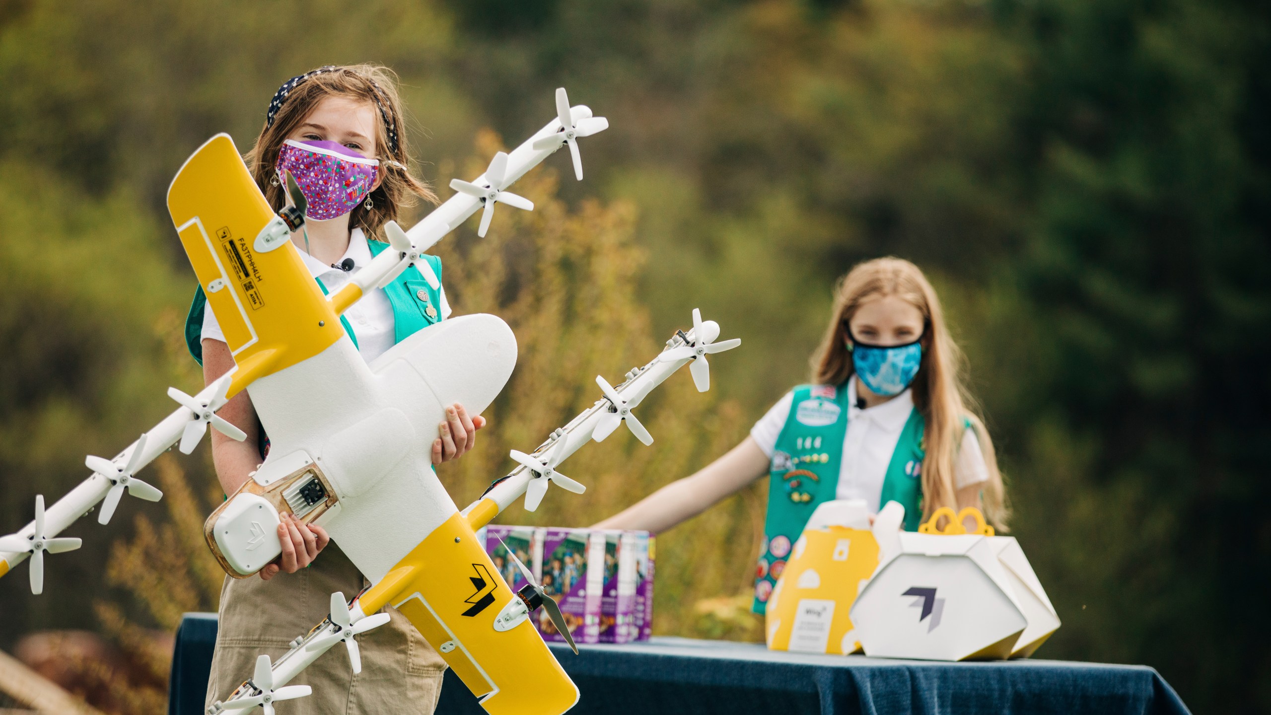 In this April 14, 2021 image provided by Wing LLC., Girl Scouts Alice Goerlich, right, and Gracie Walker pose with a Wing delivery drone in Christiansburg, Va. The company is testing drone delivery of Girl Scout cookies in the area. (Sam Dean/ Wing LLC via AP)