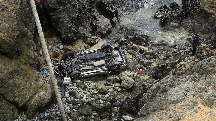 Bodega Bay firefighters work to secure the scene of a crash after a vehicle plummeted from the Bodega Head parking lot in Bodega Bay, Calif., through a wood barrier, left, landing upside down 100 feet to the rocky shoreline, killing two people in the SUV, Saturday, April 3, 2021. (Kent Porter/The Press Democrat via AP)