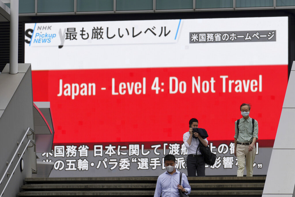 People wearing masks to help protect against the spread of the coronavirus walk in front of a screen showing the news on U.S. warning against visits to Japan Tuesday, May 25, 2021, in Tokyo. (AP Photo/Eugene Hoshiko)