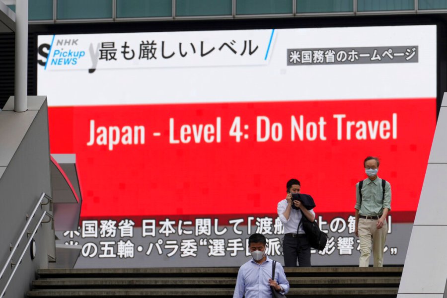 People wearing masks to help protect against the spread of the coronavirus walk in front of a screen showing the news on U.S. warning against visits to Japan Tuesday, May 25, 2021, in Tokyo. (AP Photo/Eugene Hoshiko)