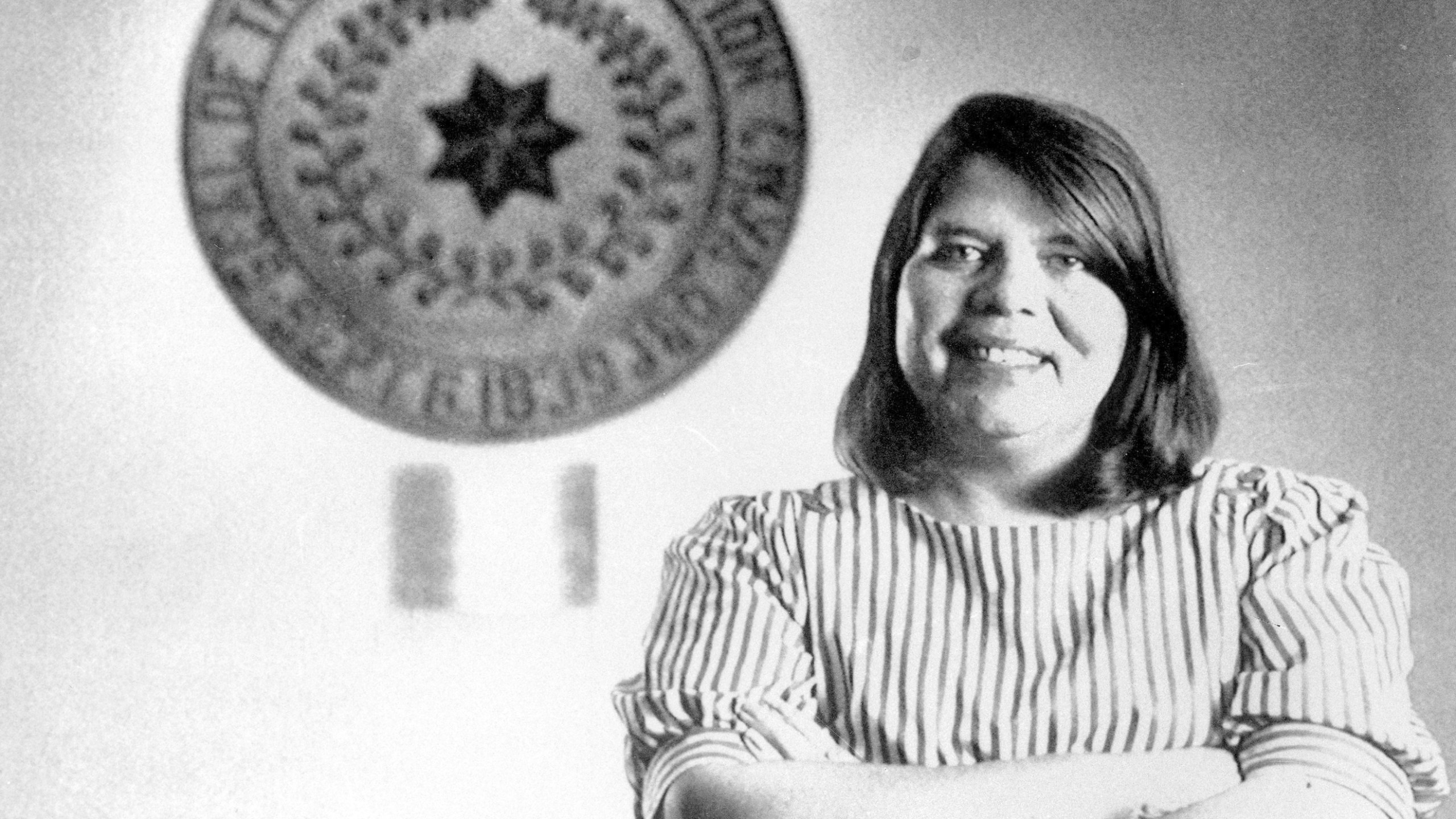 In this July 19, 1985, file photo, Wilma Mankiller, the first woman elected chief of the Cherokee Nation, poses in front of the tribal emblem at the Cherokee Nation in Oklahoma. The U.S. Mint says Mankiller will be among the first five women honored in the new American Women Quarters Program, which starts in 2022 and continues until 2025. (AP Photo, File)