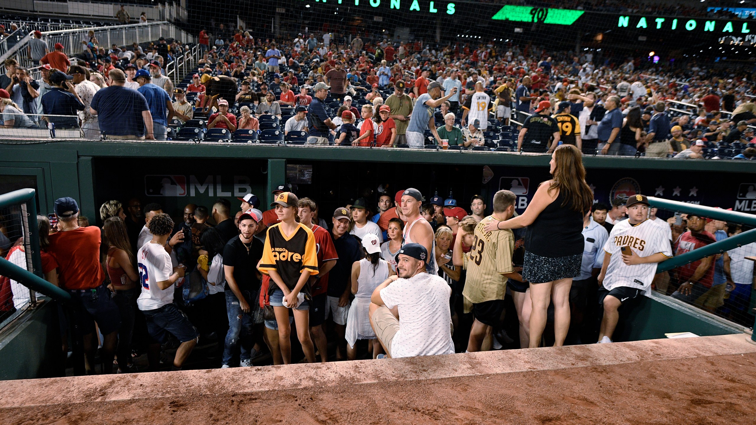 Spectators stand in the visiting team dugout during a stoppage in play due to an incident near the ballpark in the sixth inning of a baseball game between the Washington Nationals and the San Diego Padres, Saturday, July 17, 2021, in Washington. (AP Photo/Nick Wass)