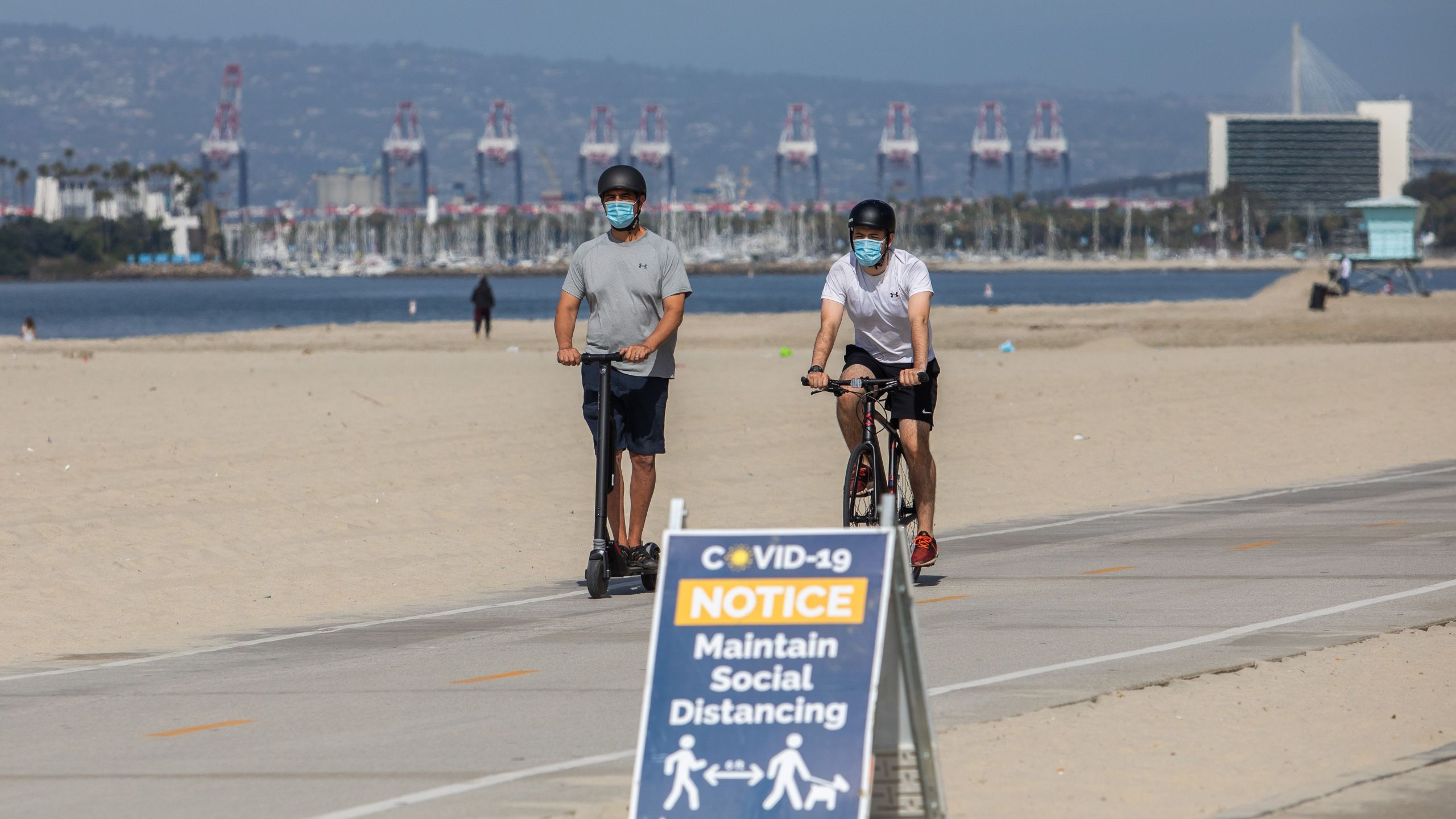 Men wearing facemasks ride a scooter and a bike near a notice about maintaining social distance on the beach in Long Beach on July 14, 2020. (APU GOMES/AFP via Getty Images)