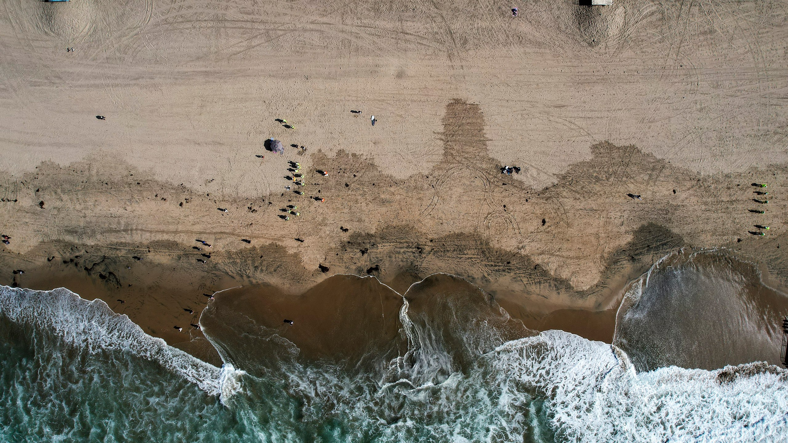 This Monday, Oct. 11, 2021, aerial file photo taken with a drone, shows beachgoers as workers in protective suits continue to clean the contaminated beach in Huntington Beach, Calif. The amount of crude oil spilled in an offshore pipeline leak in Southern California is believed to be close to about 25,000 gallons, a Coast Guard official said Thursday Oct. 14, 2021. (AP Photo/Ringo H.W. Chiu, File)