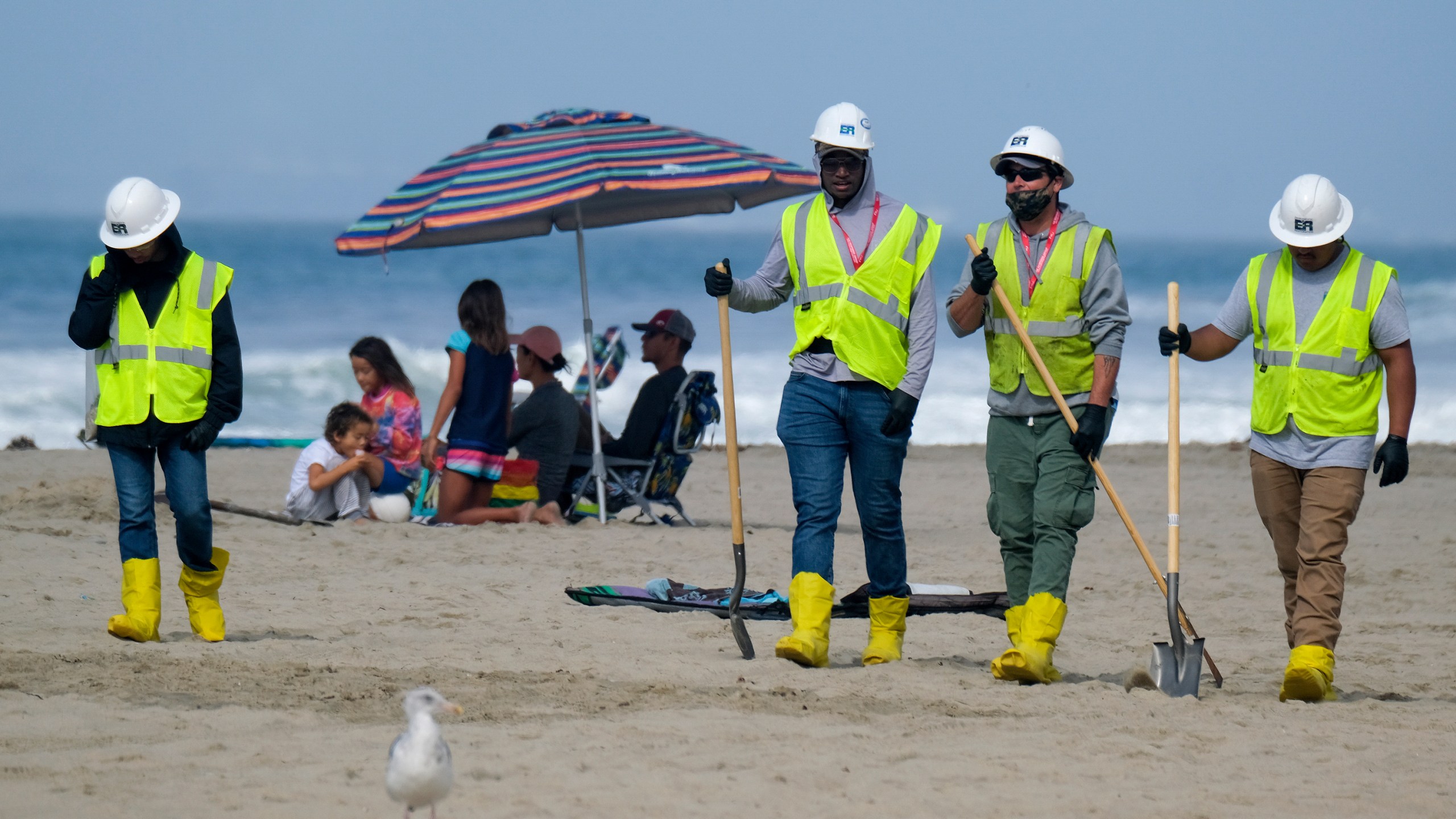 In this Oct. 11, 2021, file photo, a family, background, is under an umbrella as workers continue to clean the contaminated beach in Huntington Beach, Calif. California's uneasy relationship with the oil industry is being tested again by the latest spill to foul beaches and kill birds and fish off Orange County. (AP Photo/Ringo H.W. Chiu)