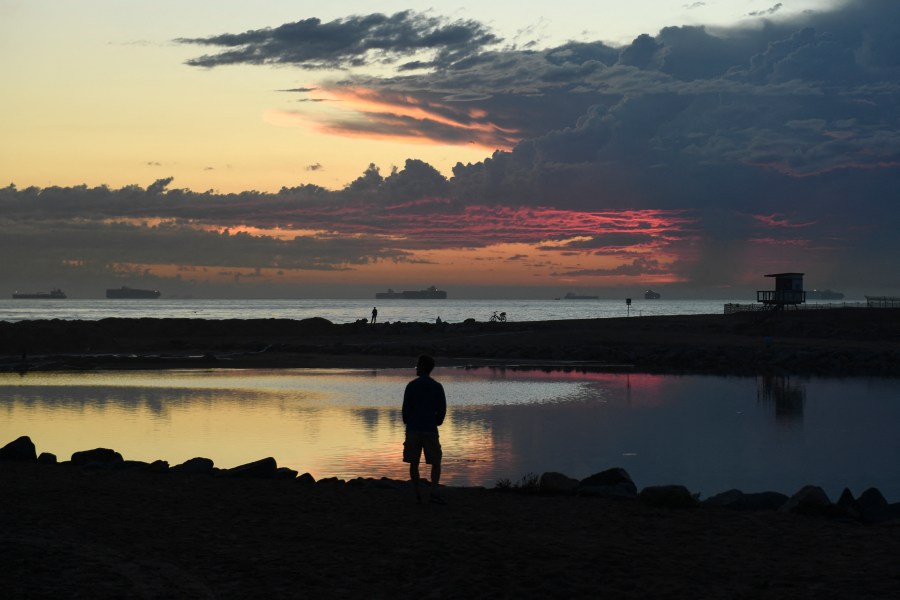 People watch the sunset from an inlet to the Santa Ana River blocked with sand and barriers to protect the inflow of oil as cargo container ships wait off the coast to enter the Port of Long Beach and Port of Los Angeles after an oil spill in the Pacific Ocean as seen from Huntington Beach, California on Oct. 4, 2021. (PATRICK T. FALLON/AFP via Getty Images)