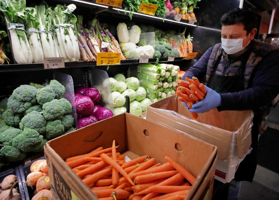 In this March 27, 2020, file photo, a worker, wearing a protective mask against the coronavirus, stocks produce before the opening of Gus's Community Market in San Francisco. (AP Photo/Ben Margot, File)