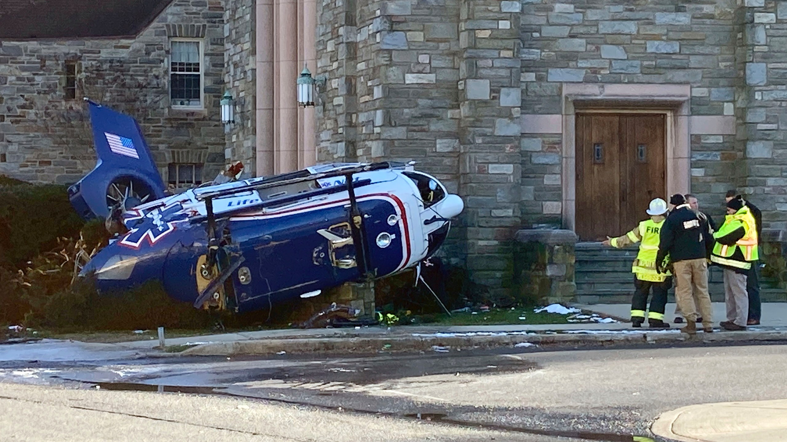 Firefighters from Upper Darby, Pa, stand near a medical helicopter that crashed next to a church in a residential area of suburban Philadelphia, Tuesday, Jan. 11, 2022, with four people on board including an infant. All are expected to survive, authorities said. (AP Photo/Claudia Lauer)