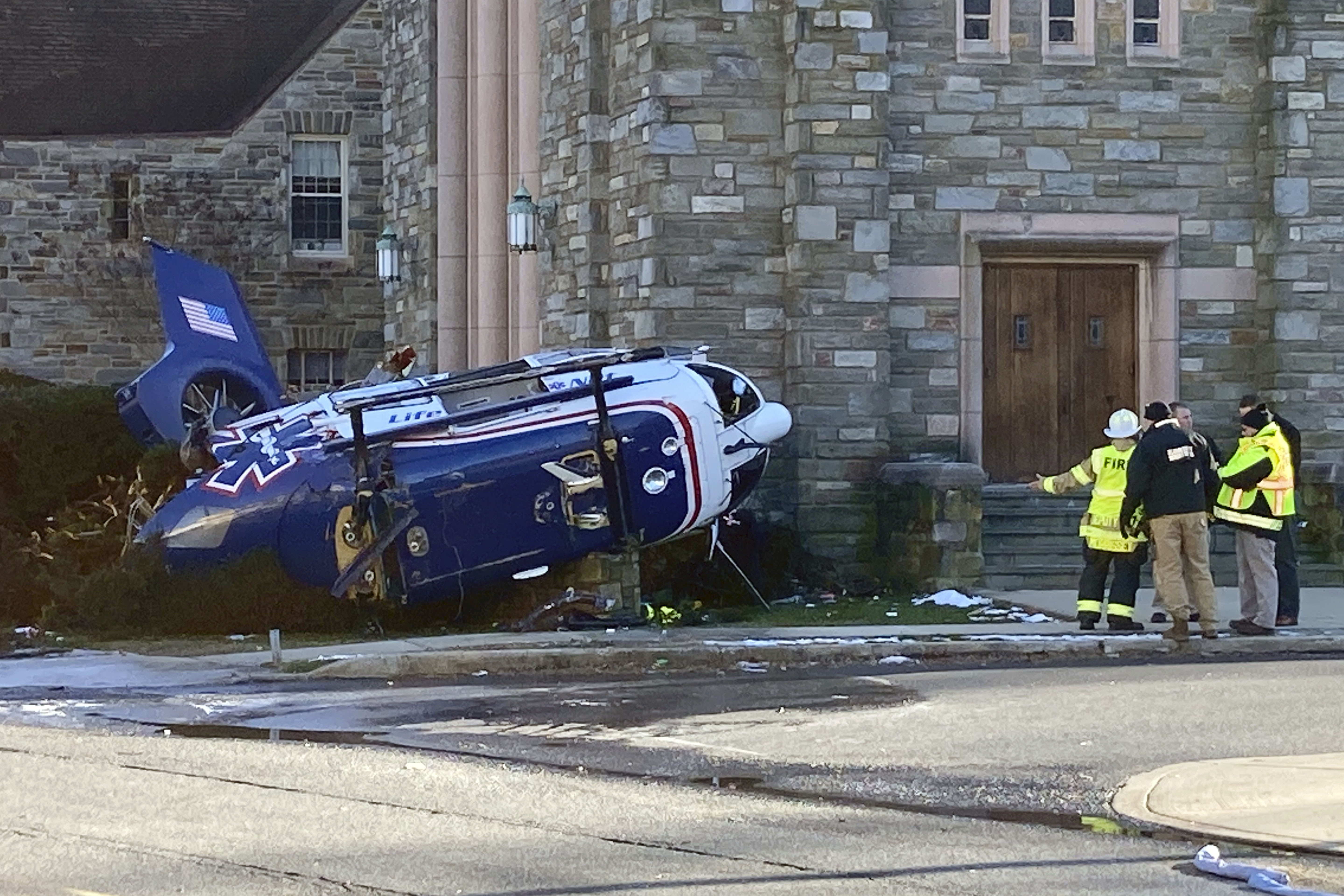 Firefighters from Upper Darby, Pa, stand near a medical helicopter that crashed next to a church in a residential area of suburban Philadelphia, Tuesday, Jan. 11, 2022, with four people on board including an infant. All are expected to survive, authorities said. (AP Photo/Claudia Lauer)