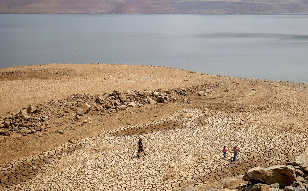 In this Aug. 22, 2021, file photo, a family walks over cracked mud near Lake Oroville's shore as water levels remain low due to continuing drought conditions in Oroville, Calif. (AP Photo/Ethan Swope, File)