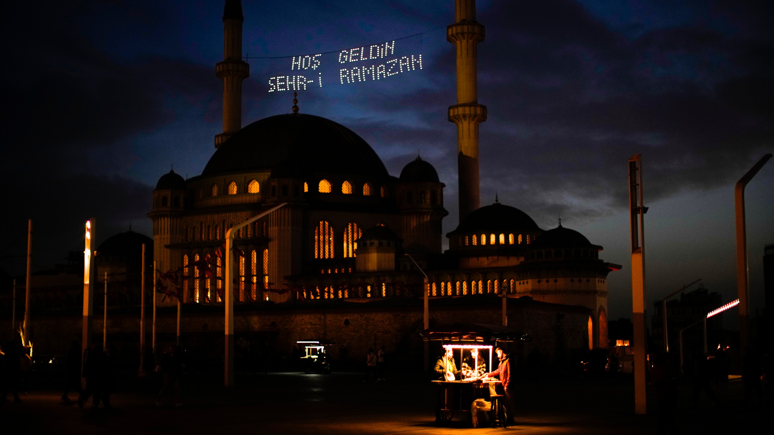 People buy roasted chestnuts next to Taksim mosque as the sun sets during the first day of the Muslim holy fasting month of Ramadan, in Istanbul, Turkey, Saturday, April 2, 2022. Lights on top read in Turkish: "Welcome Sehr-i Ramadan". (AP Photo/Francisco Seco)