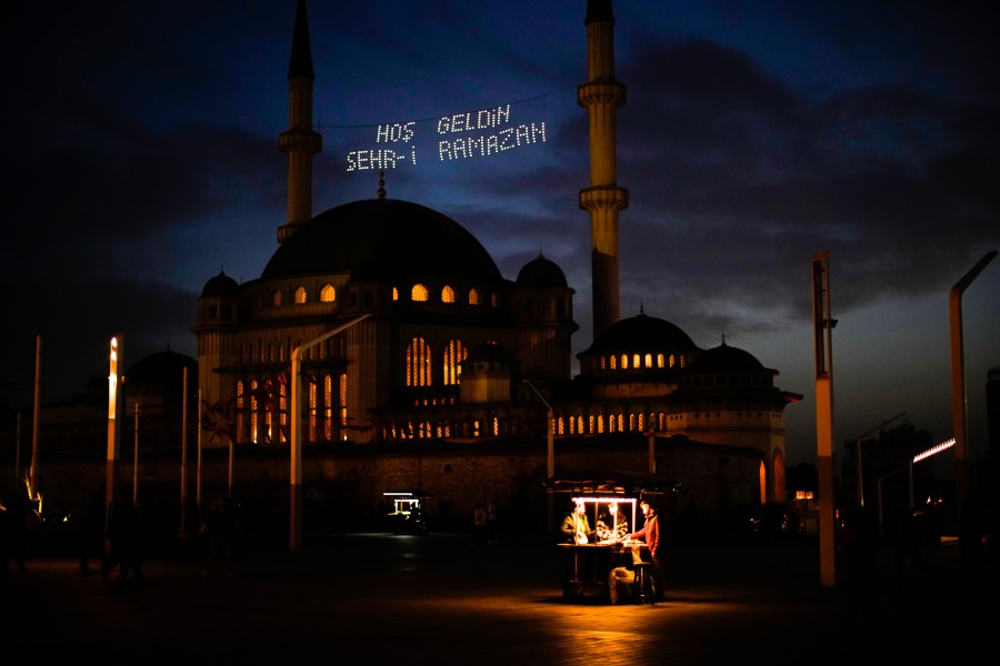 People buy roasted chestnuts next to Taksim mosque as the sun sets during the first day of the Muslim holy fasting month of Ramadan, in Istanbul, Turkey, Saturday, April 2, 2022. Lights on top read in Turkish: "Welcome Sehr-i Ramadan". (AP Photo/Francisco Seco)