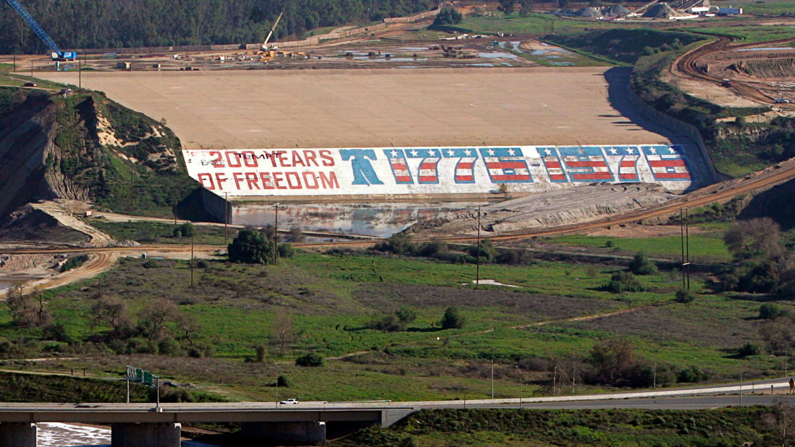 Seen is the Prado Dam with its bicentennial mural at a time when crews were working to close a breach in the dam, at left, due to heavy rains in Corona, Calif., on Jan. 14, 2005. A giant mural that students painted on a Southern California dam for the U.S. bicentennial in 1976 is being recreated after years of controversy over removal of the original due to environmental concerns. Workers following the original design began painting the new mural on the spillway of Prado Dam in April 2023, Southern California News Group reported. (AP Photo/Chris Carlson, File)