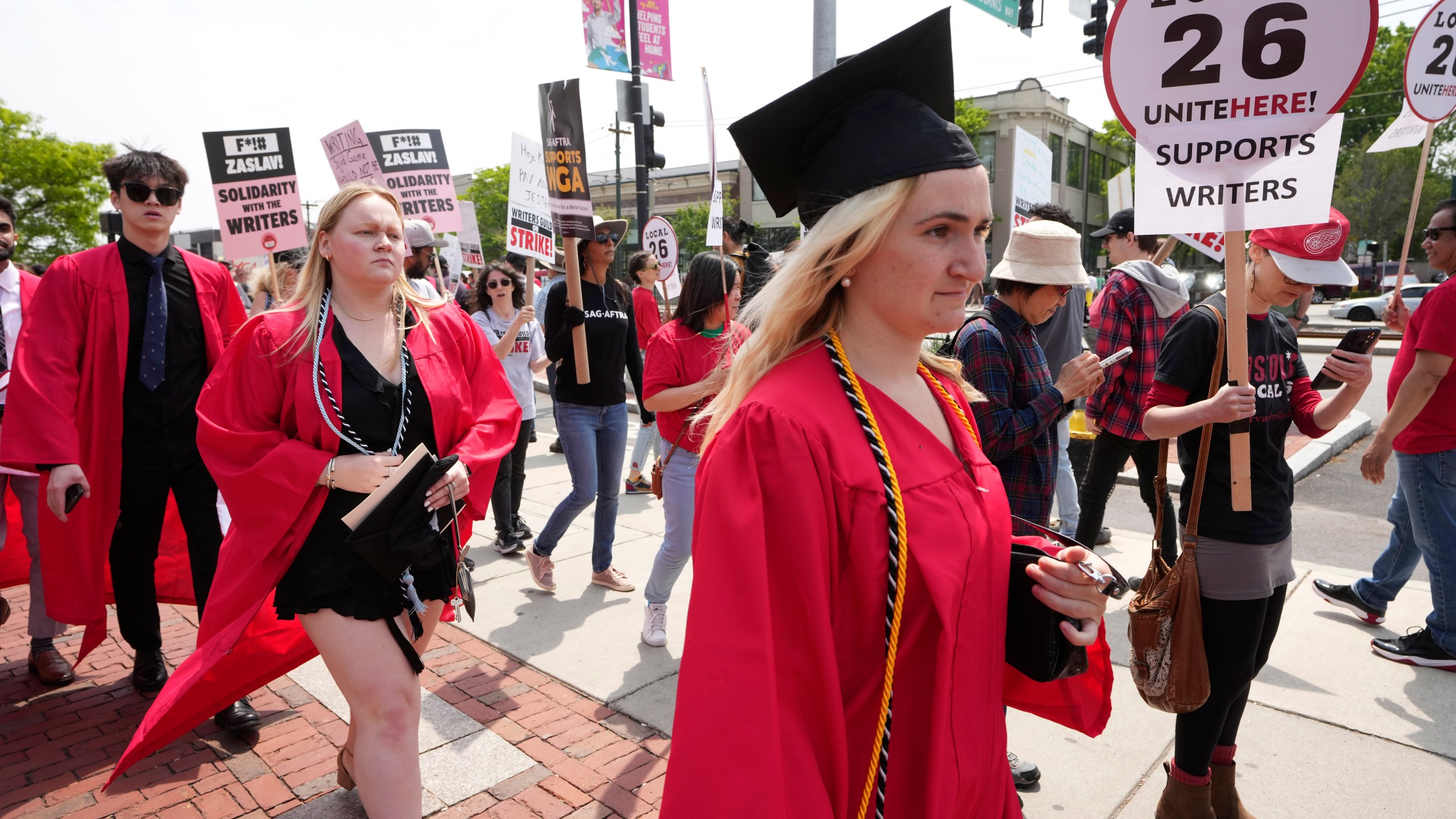 People dressed in commencement gowns, front, walk past protesters supporting the Hollywood writers' strike outside an entrance to Boston University commencement ceremonies, Sunday, May 21, 2023, in Boston. David Zaslav, president and CEO of Warner Bros. Discovery, delivered an address during the ceremonies Sunday. (AP Photo/Steven Senne)