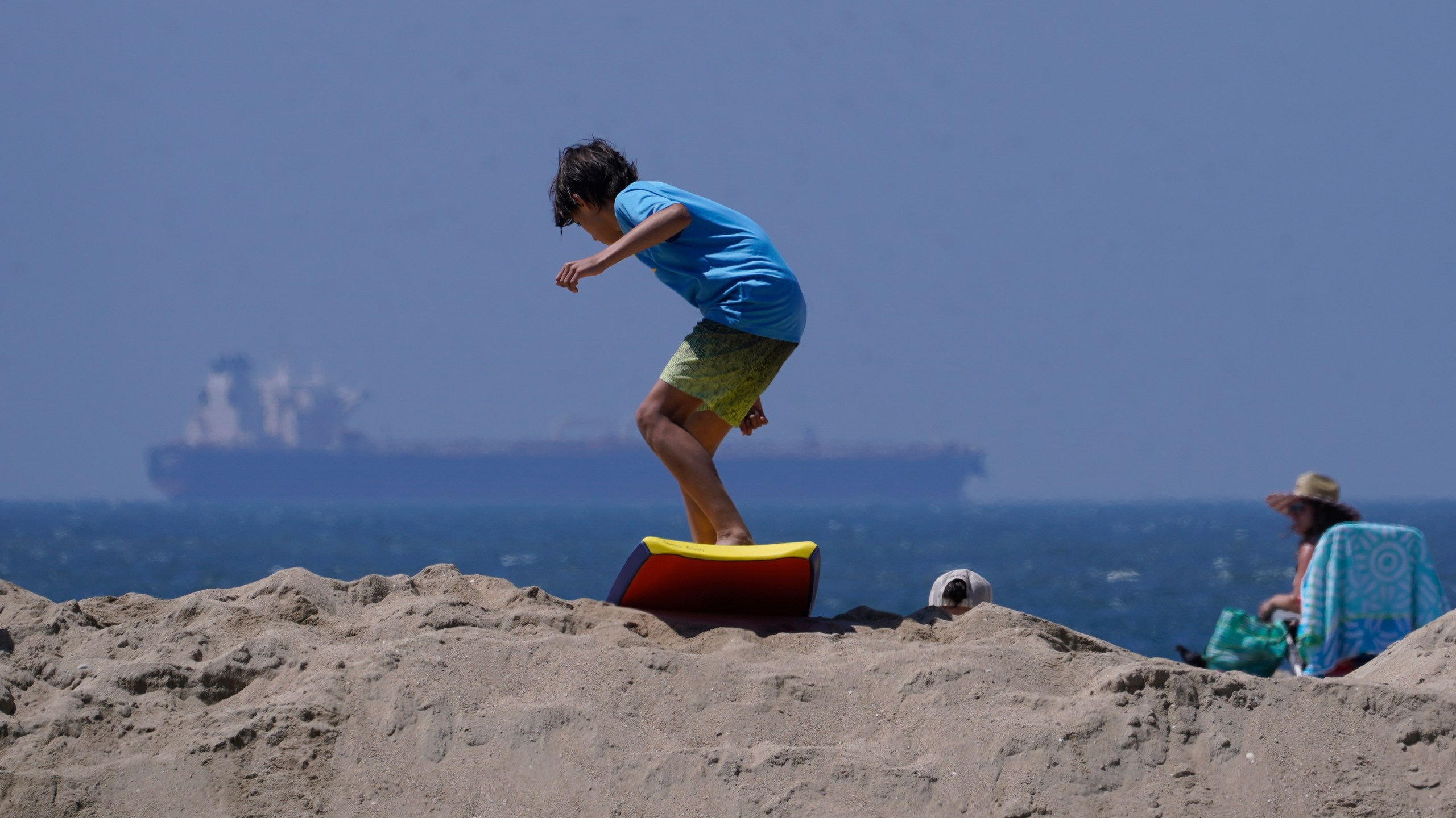 A boy slides with a surf board on top of a sand berm in Seal Beach, Calif., Friday, Aug. 18, 2023. Officials in Southern California were also re-enforcing sand berms, built to protect low-lying coastal communities against winter surf. Hurricane Hilary is churning off Mexico's Pacific coast as a powerful Category 4 storm threatening to unleash torrential rains on the mudslide-prone border city of Tijuana before heading into Southern California as the first tropical storm there in 84 years. (AP Photo/Damian Dovarganes)