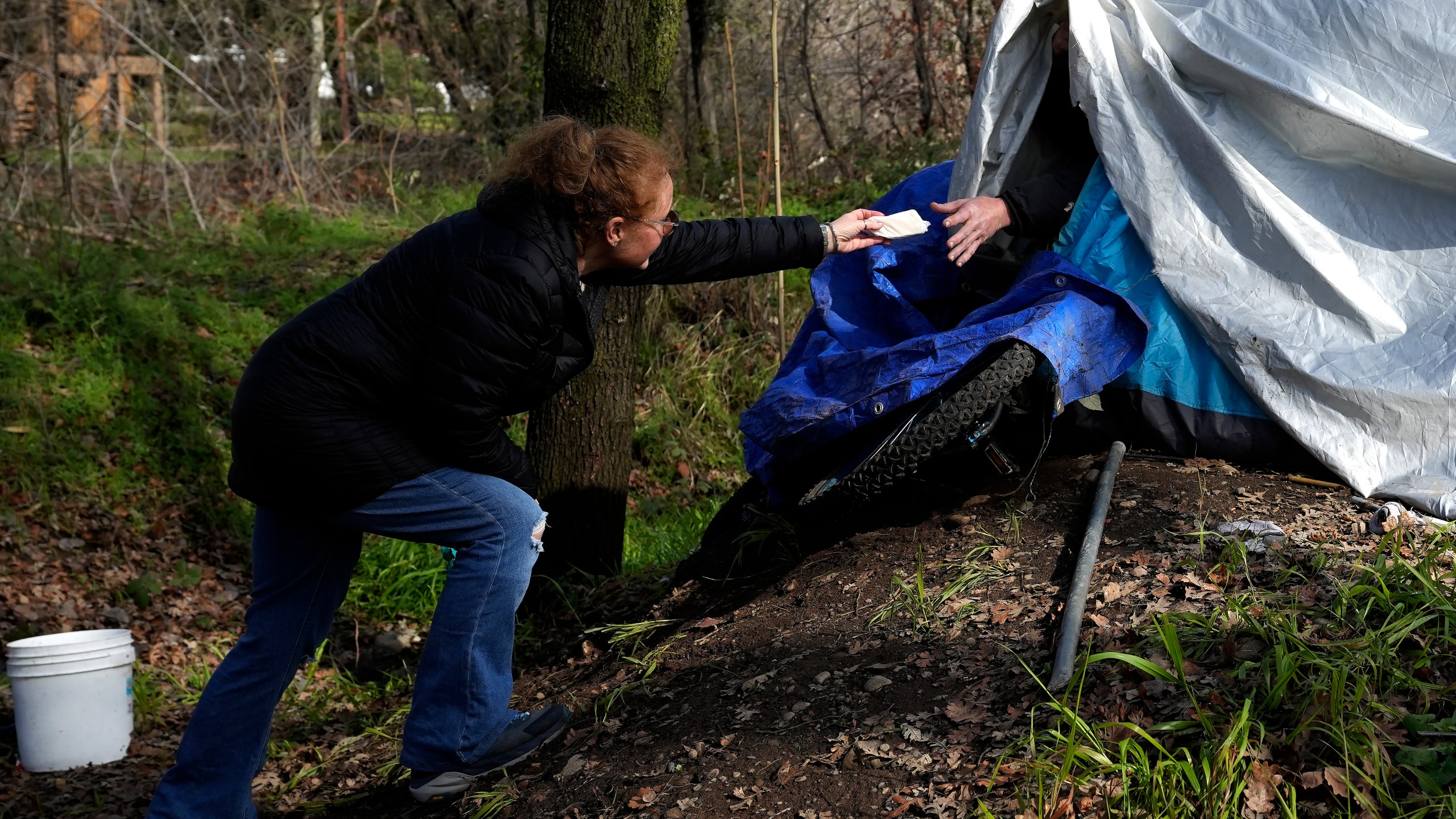 Resiliency Empowerment Support Team (REST) team member Beth Perkins, give a homeless person a hand warmer during a visit to a homeless camp in Chico, Calif., Feb. 8, 2024. A measure aimed at transforming how California spends money on mental health will go before voters in March as the state continues to grapple an unabated homelessness crisis. The REST Program does daily visits to homeless encampments to get them into treatment or housing. Butte County officials fear the REST program would lose its funding if California voters approve Proposition 1.(AP Photo/Rich Pedroncelli)