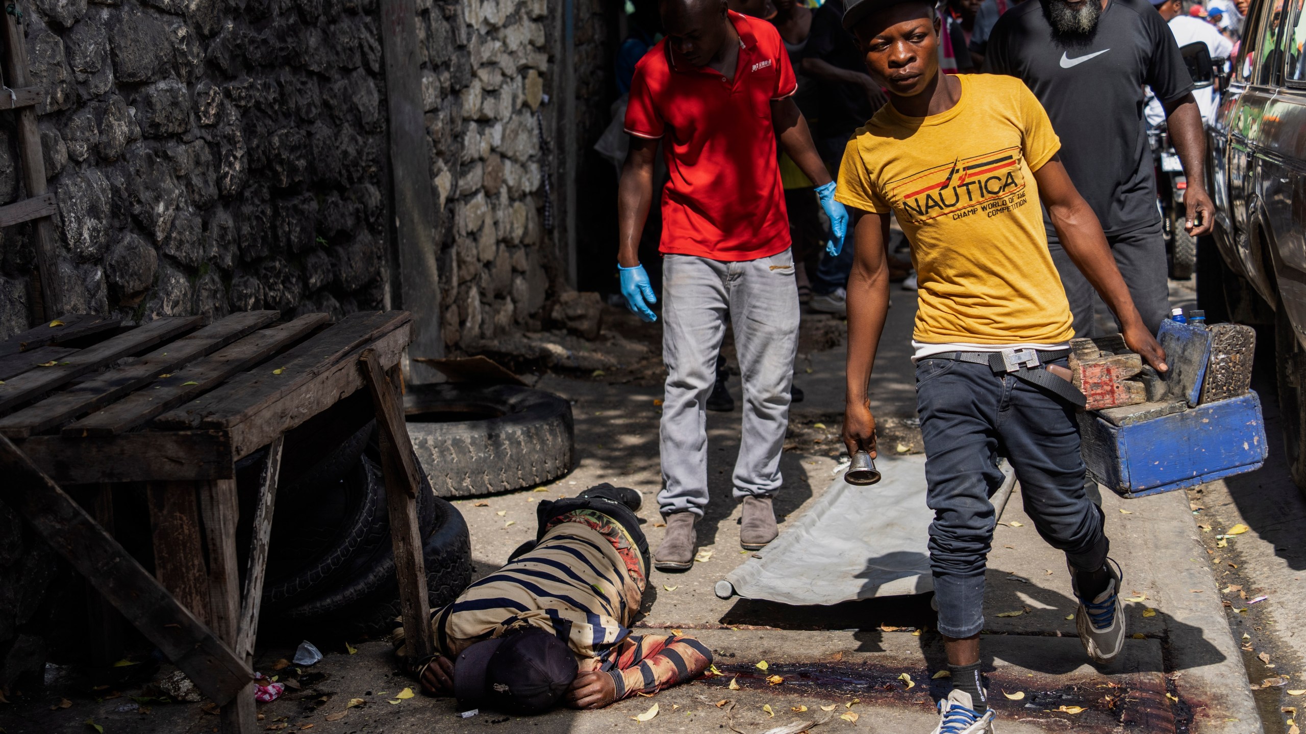 A shoeshiner walks past a bloodied body being removed by forensic workers in Port-au-Prince, Haiti, Wednesday, May 1, 2024. Forensics said the body had gunshot wounds. (AP Photo/Ramon Espinosa)