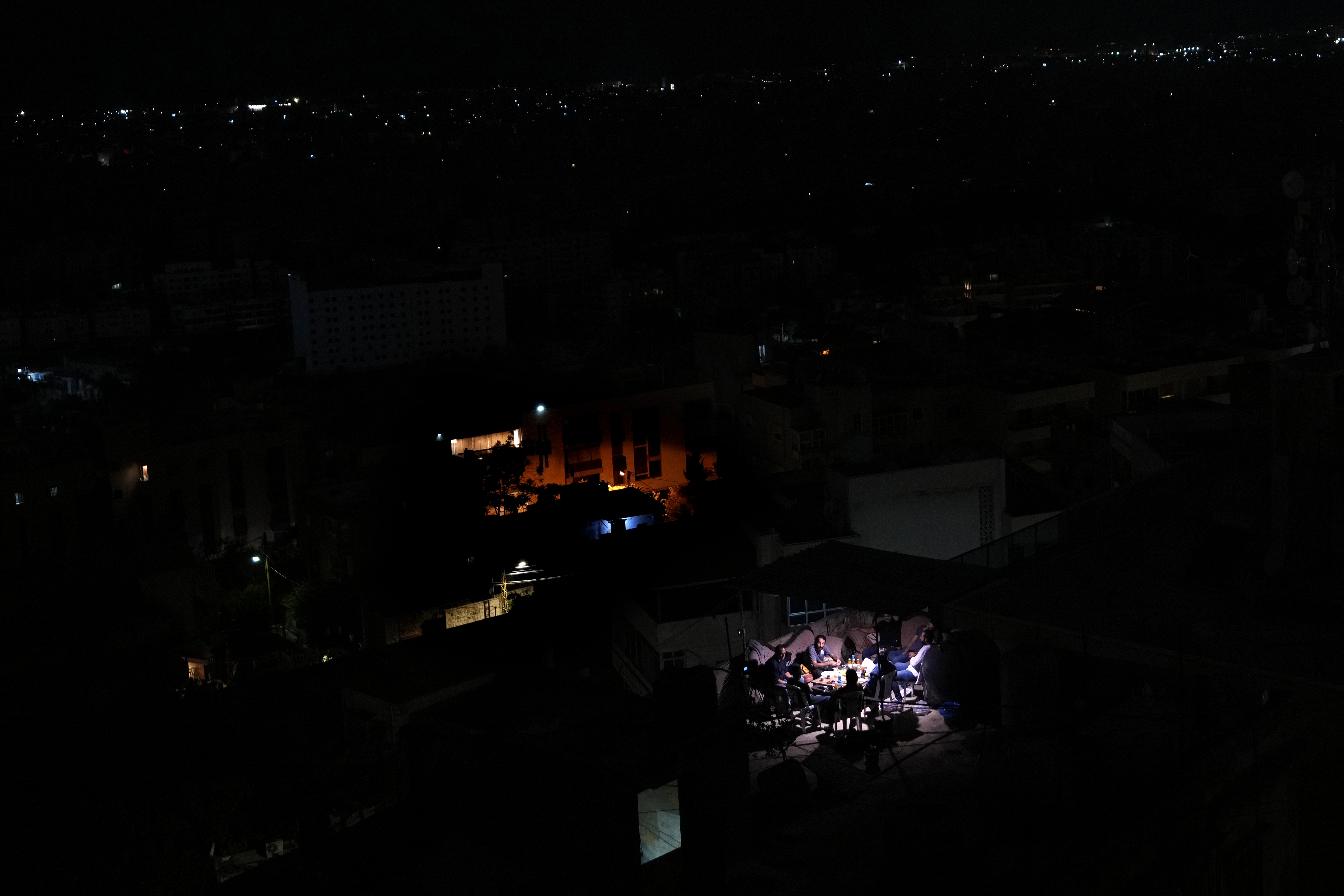 Residents sit on the roof of a building and have dinner as Dahiyeh suburb, background, remains in darkness after Israeli airstrikes, Lebanon, Friday, Oct. 11, 2024. (AP Photo/Hassan Ammar)