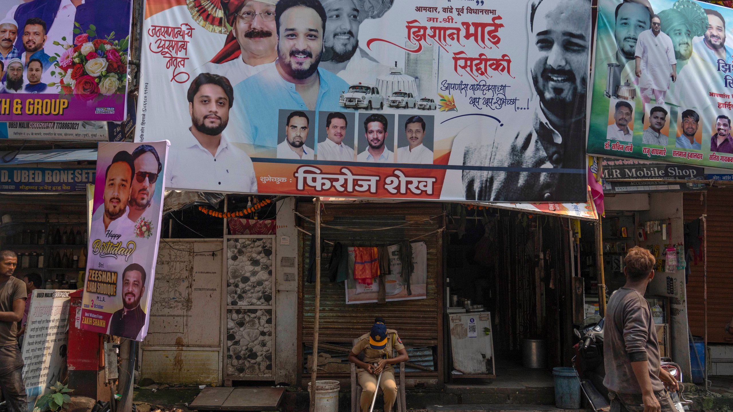 A police person sits below a large hoarding showing a portrait in red turban of Baba Siddique, a senior politician, at the spot where Siddique was shot at outside his son's office in Mumbai and later succumbed to his wounds in a hospital on Saturday night, in Mumbai, India, Sunday, Oct. 13, 2024. (AP Photo/Rafiq Maqbool)