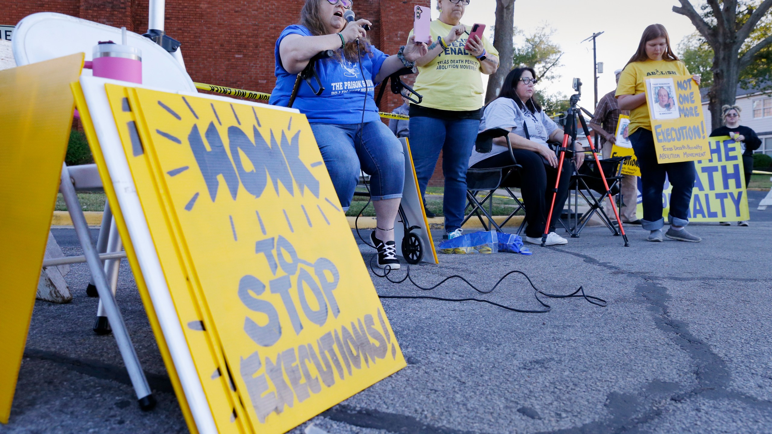 Dani Allen, center left with microphone, an anti-death penalty advocate, speaks during a protest outside the prison where Robert Roberson is scheduled for execution at the Huntsville Unit of the Texas State Penitentiary, Thursday, Oct. 17, 2024, in Huntsville, Texas. (AP Photo/Michael Wyke)