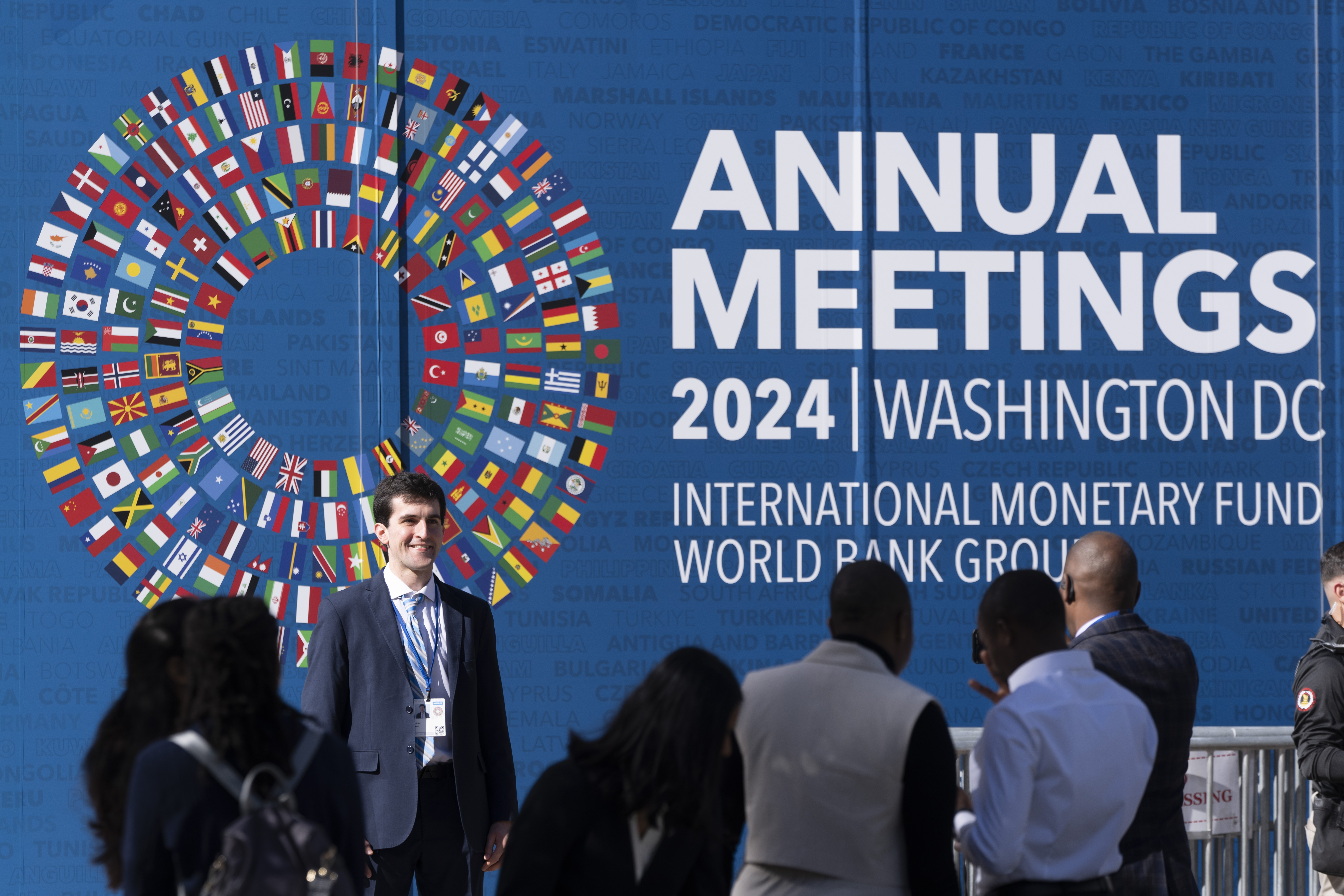 An attendee poses for a photograph in front of a sign advertising the annual meetings of the International Monetary Fund (IMF) and the World Bank Group (WBG), which started in Washington on Monday, Oct. 21, 2024. (AP Photo/Ben Curtis)