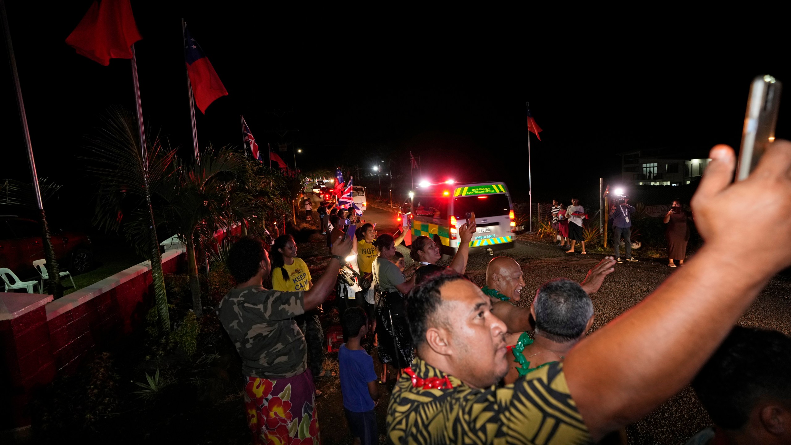 People wave and take pictures as the motorcade with Britain's King Charles III and Queen Camilla passes in the village of Siumu, Samoa, on Wednesday, Oct. 23, 2024. (AP Photo/Rick Rycroft)