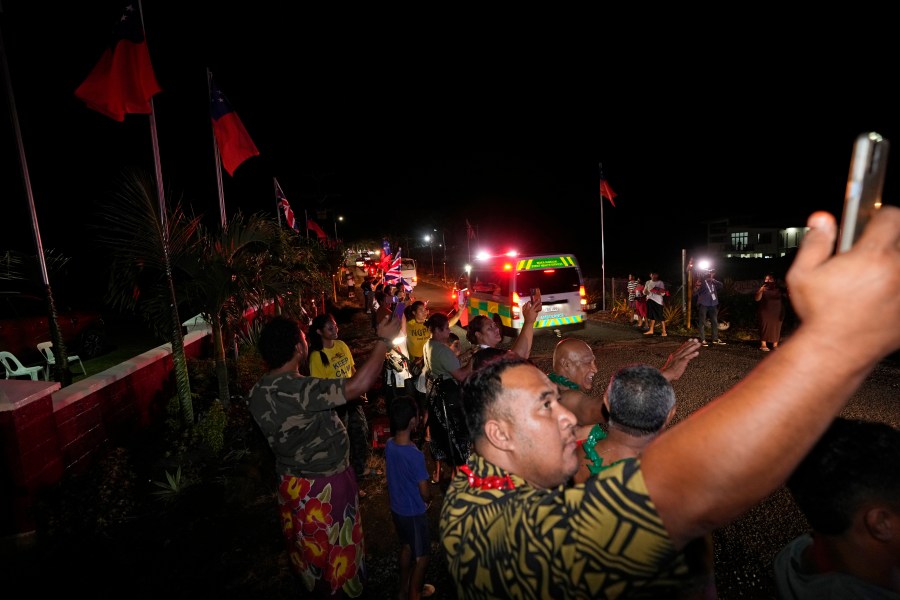 People wave and take pictures as the motorcade with Britain's King Charles III and Queen Camilla passes in the village of Siumu, Samoa, on Wednesday, Oct. 23, 2024. (AP Photo/Rick Rycroft)