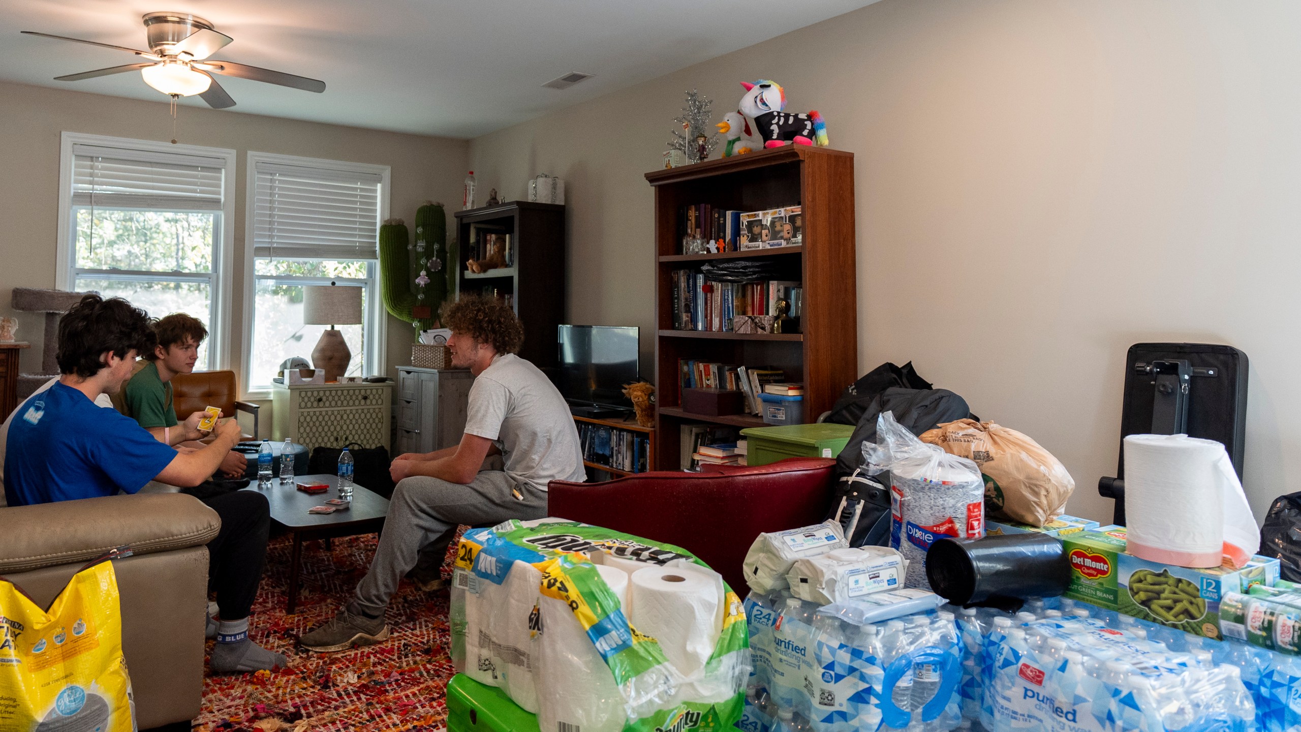 Cases of water, canned food and other supplies are seen in the living room while high school senior Ari Cohen, right, and his friends play a game of Uno, Friday, Oct. 18, 2024, in Asheville, N.C. (AP Photo/Stephanie Scarbrough)