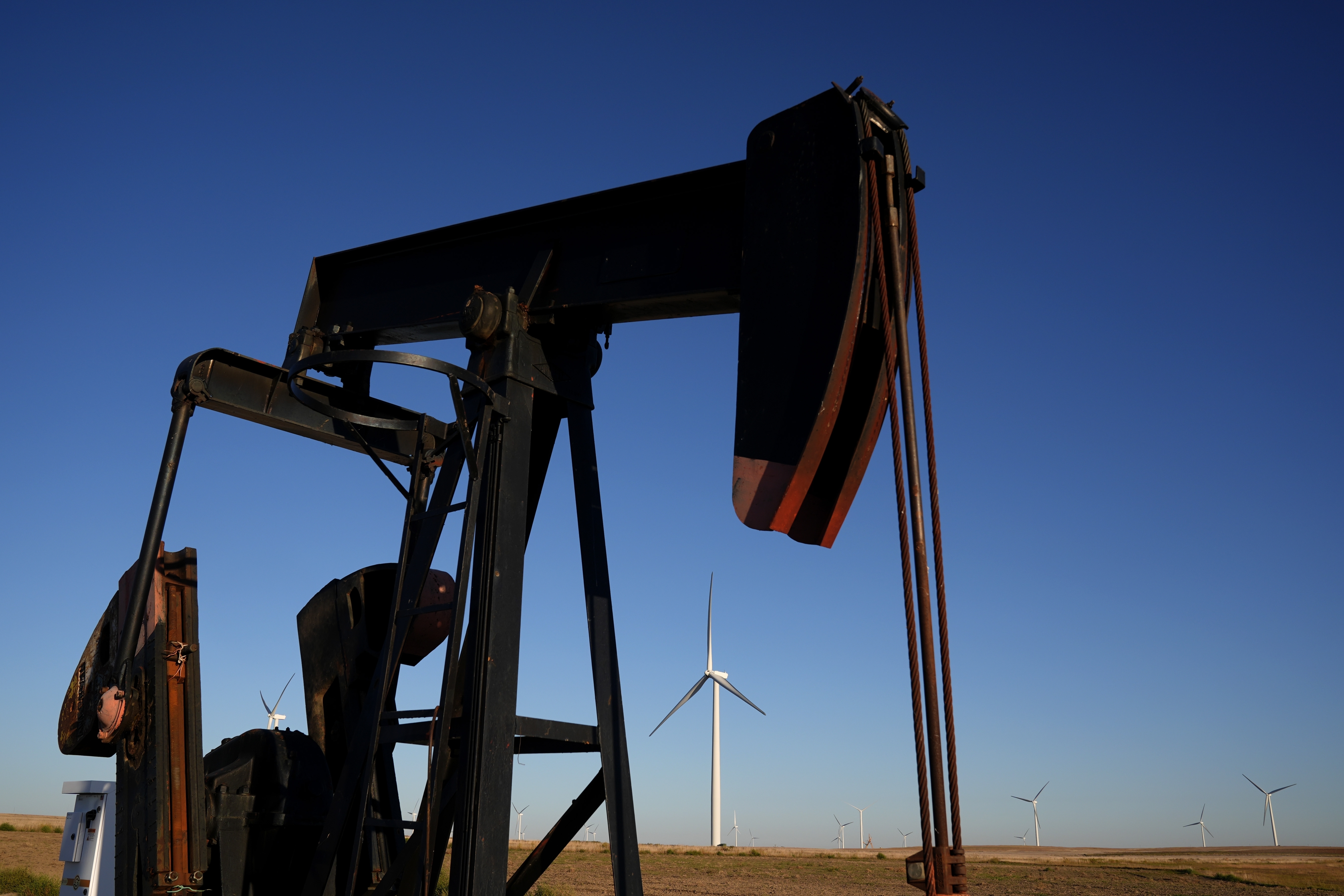 A pumpjack operates in the foreground as wind turbines at the Buckeye Wind Energy wind farm rise in the distance, Monday, Sept. 30, 2024, near Hays, Kan. (AP Photo/Charlie Riedel)