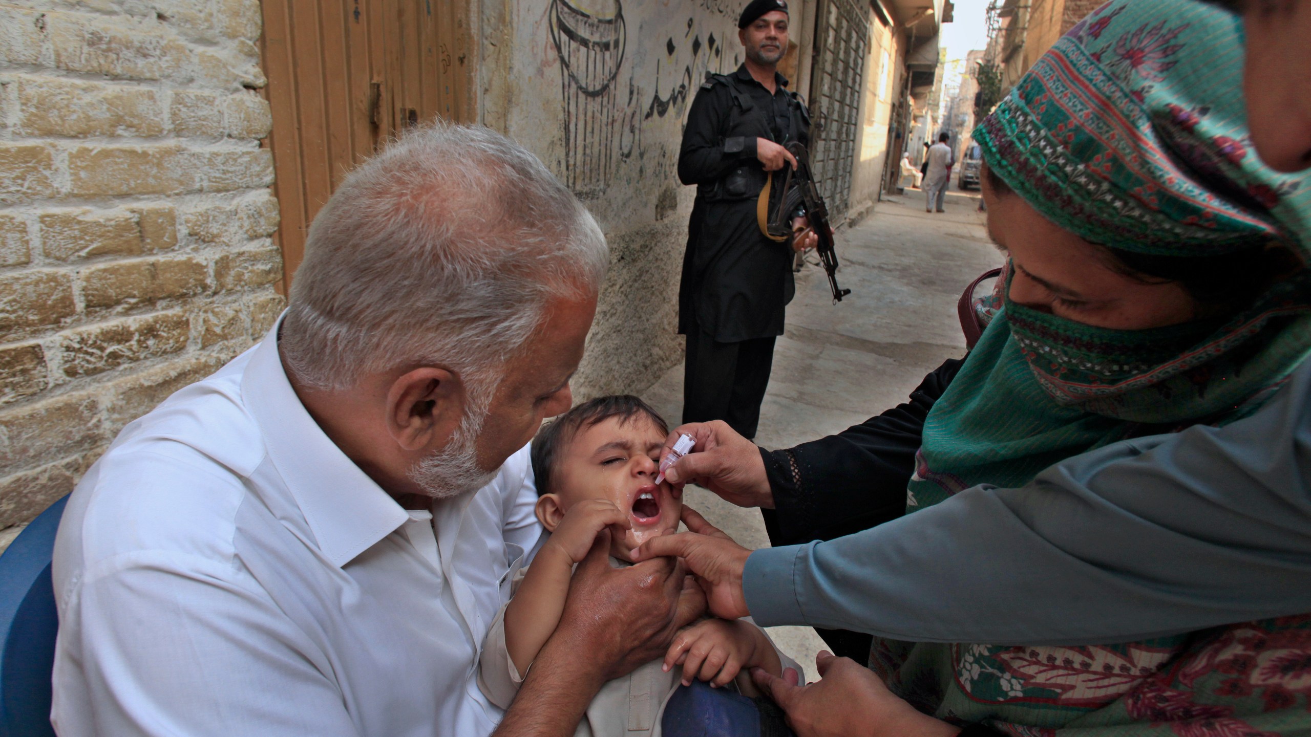 A police officer stands guard as a health worker, right, administers a polio vaccine to a child in a neighbourhood of Peshawar, Pakistan, Monday, Oct. 28, 2024. (AP Photo/Mohammad Sajjad)