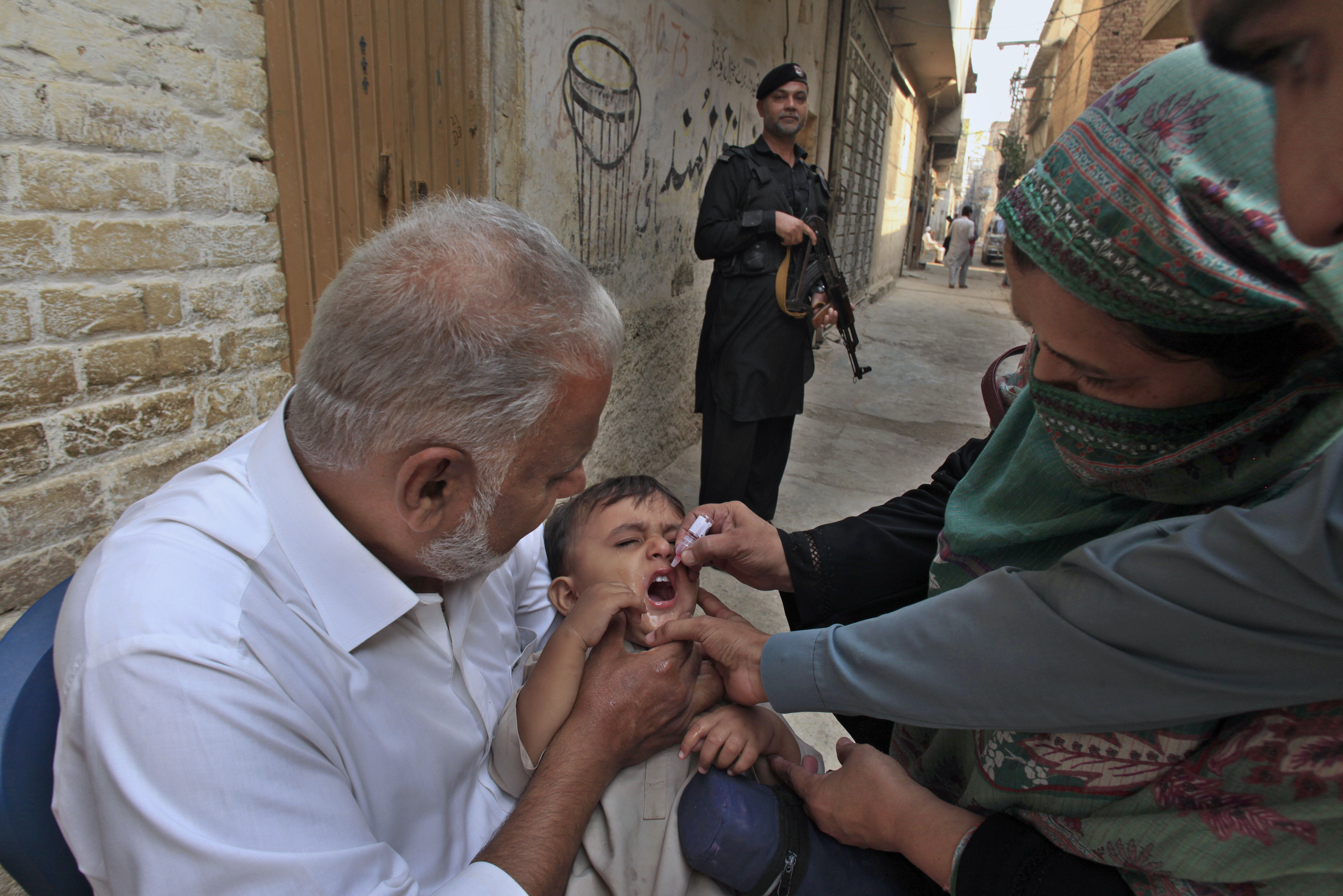 A police officer stands guard as a health worker, right, administers a polio vaccine to a child in a neighbourhood of Peshawar, Pakistan, Monday, Oct. 28, 2024. (AP Photo/Mohammad Sajjad)